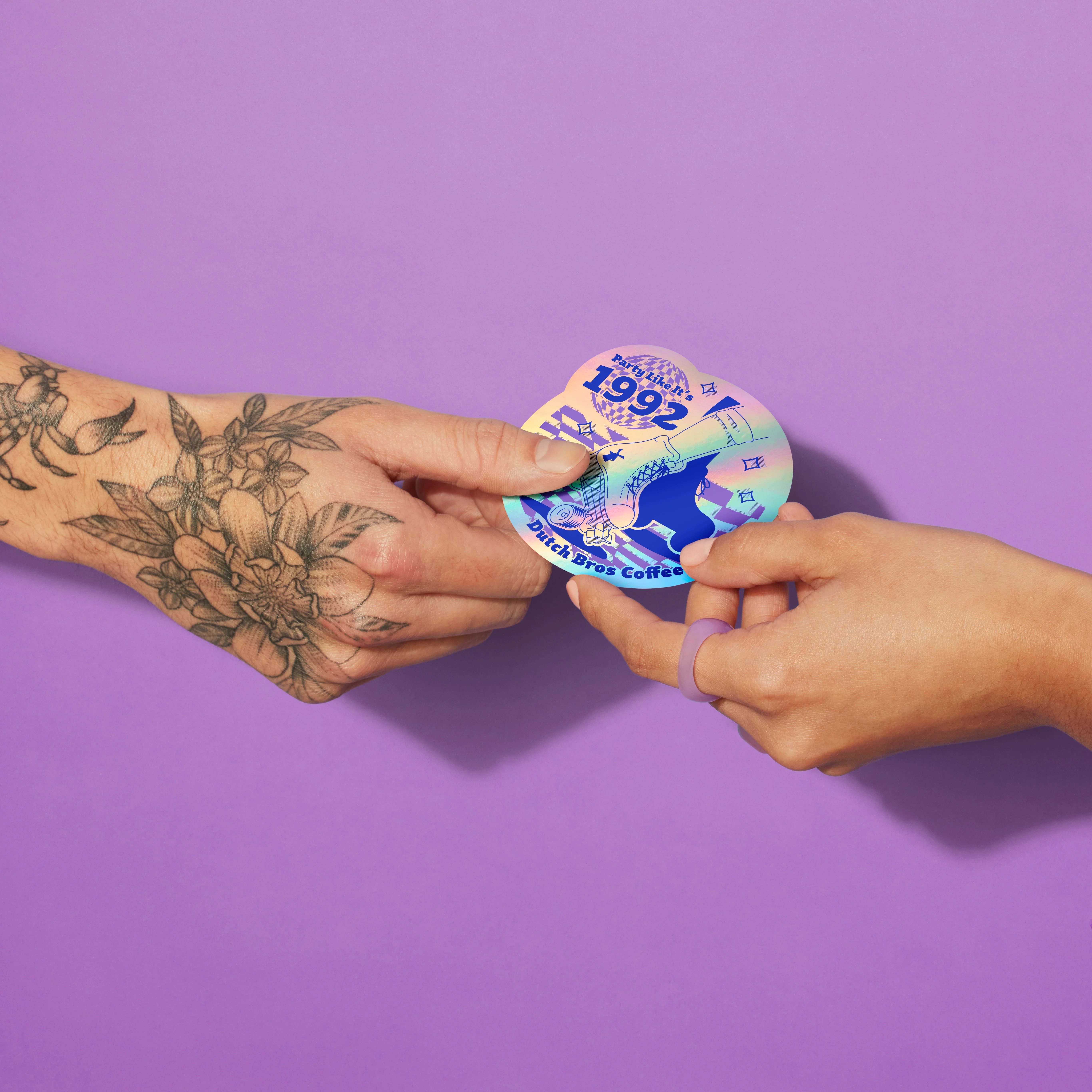 Tattooed hand passing a holographic "Party Like It's 1992" Dutch Bros Coffee sticker to another hand against purple background.