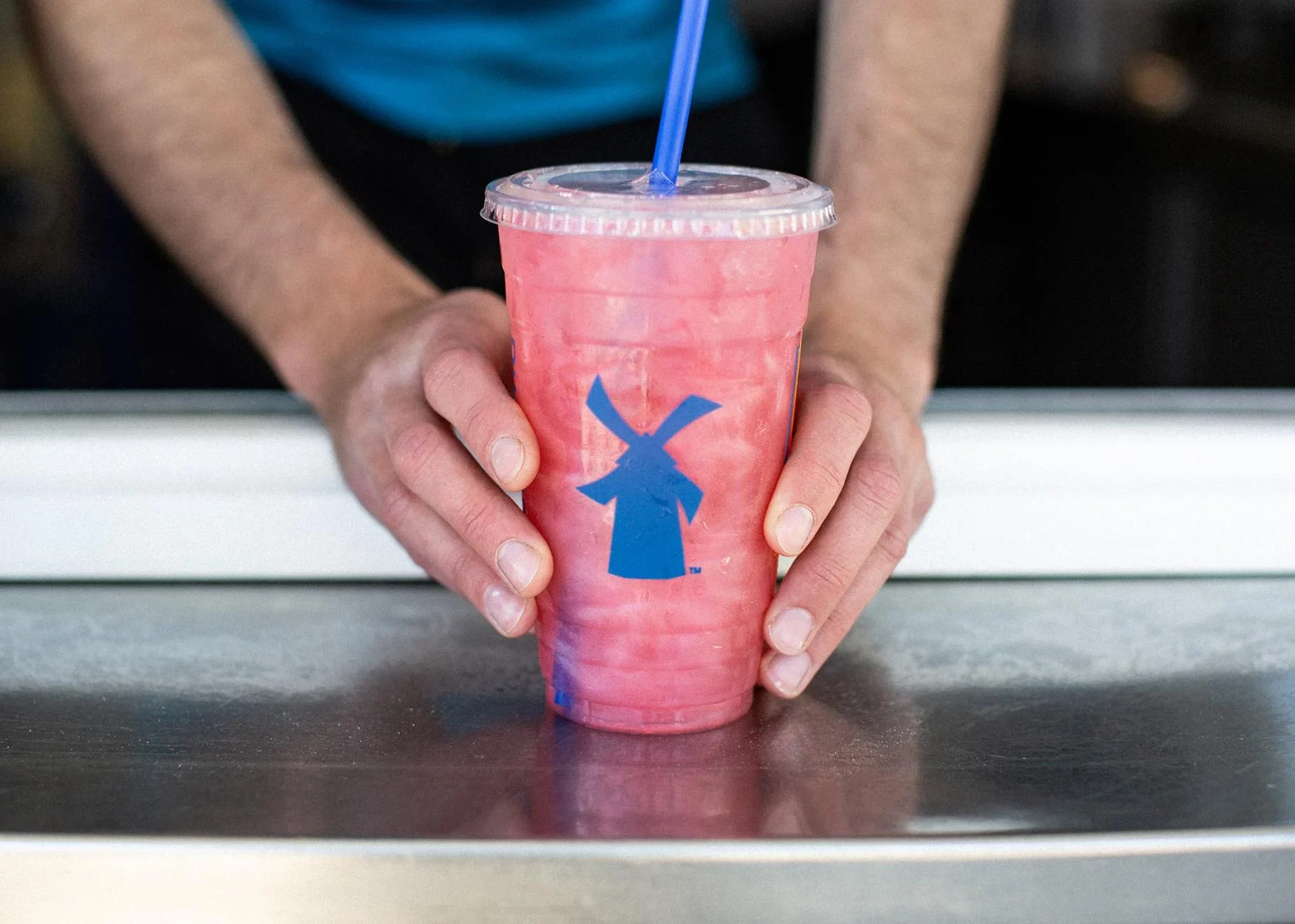 Hands holding a pink frozen drink in a clear plastic cup with blue windmill logo and straw on a counter surface.