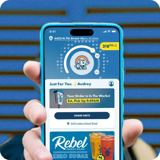 Hand holding a blue smartphone displaying the Dutch Bros app with an active order status and 218 reward points on screen.