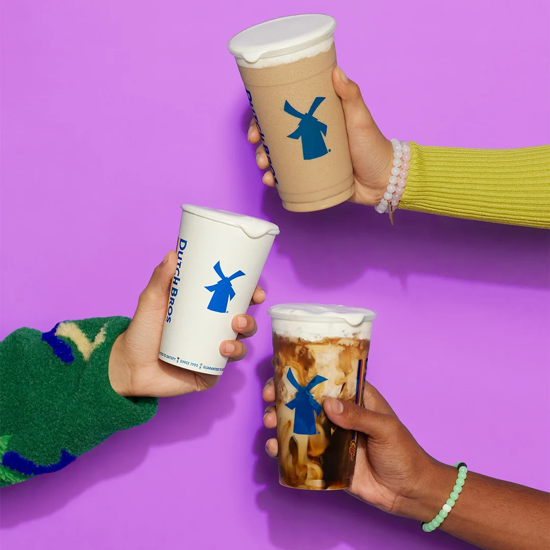 Three hands holding different Dutch Bros coffee drinks with blue windmill logos against a purple background.