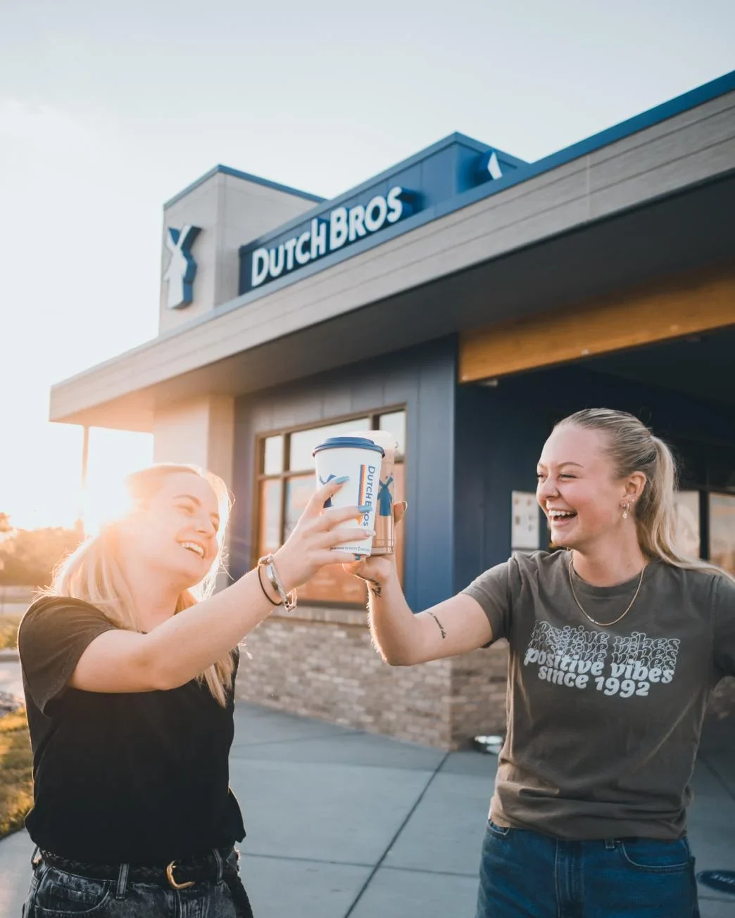 Two friends toasting Dutch Bros coffee cups outside the storefront during sunset, laughing joyfully together.