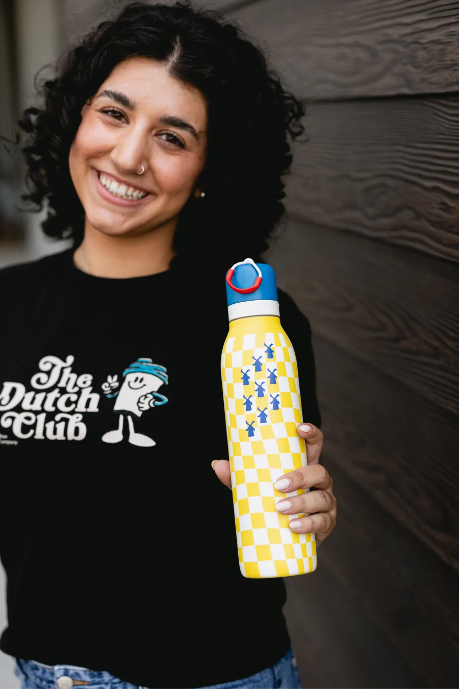 Person in "Dutch Club" shirt holding yellow and white checkered water bottle with blue windmill designs against wooden wall
