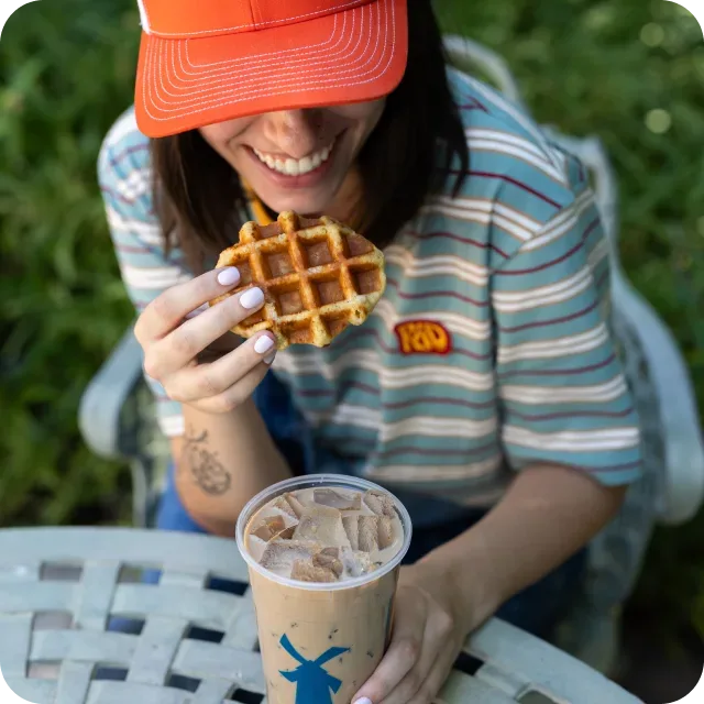 Smiling woman in an orange cap holding a waffle, with an iced coffee from Dutch Bros on an outdoor table.
