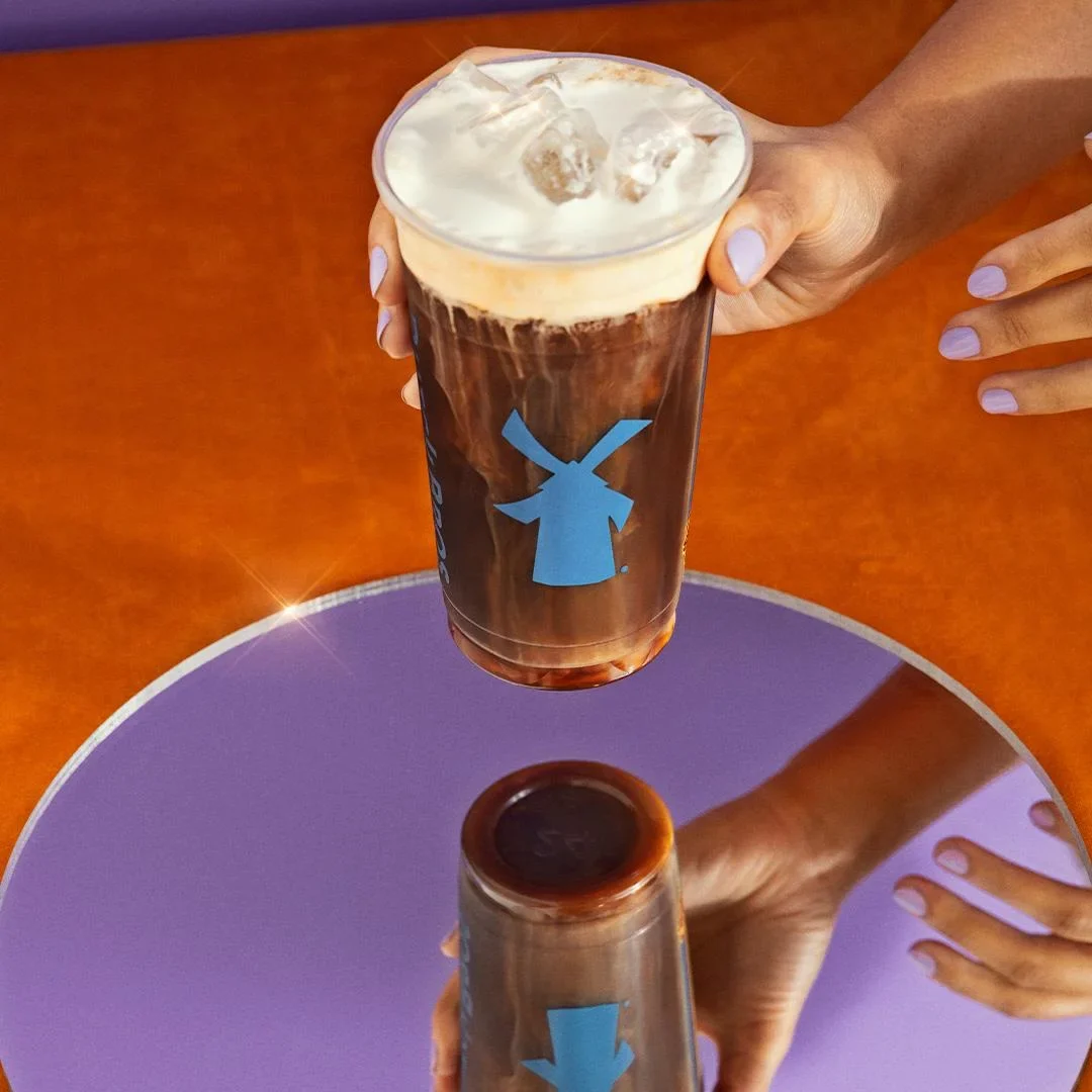 Hand holding iced coffee drink with blue windmill logo on cup, positioned above purple circular table with another drink.