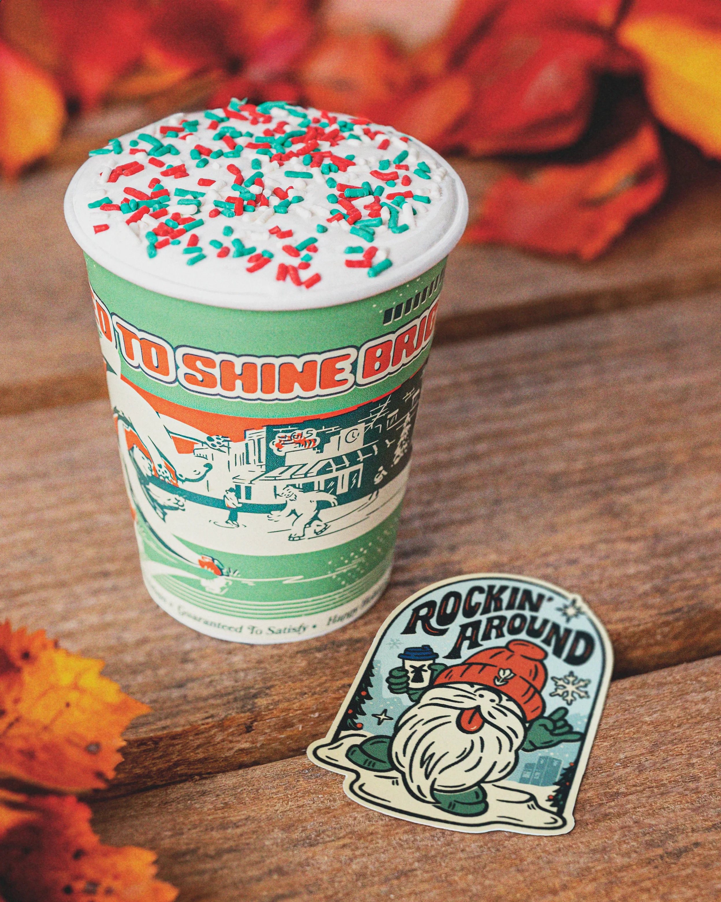 Moonshine Brew coffee cup with festive red and green sprinkles beside a Rockin' Around sticker with winter gnome design.