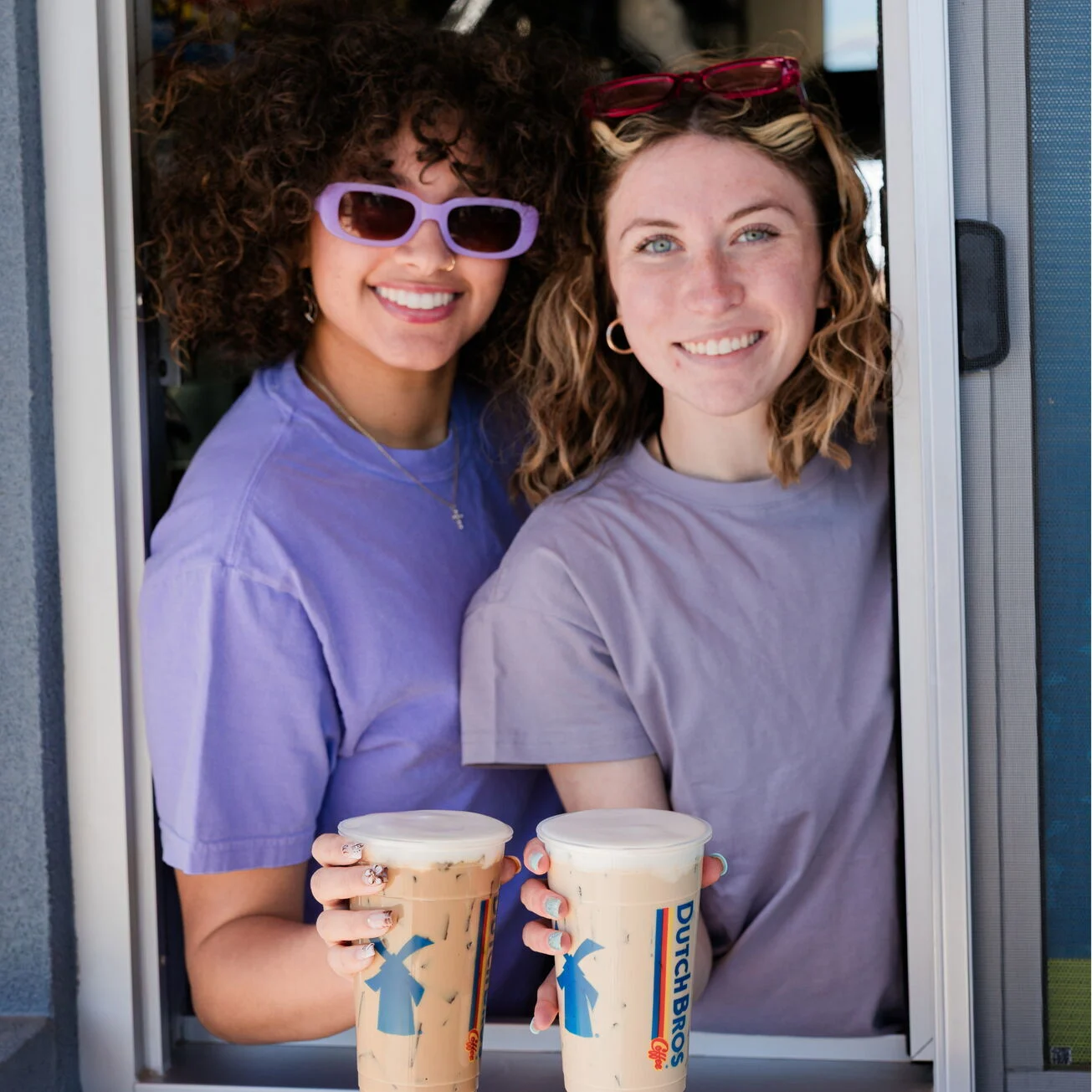 Two smiling women in purple shirts holding Dutch Bros iced coffee drinks, one wearing purple sunglasses.