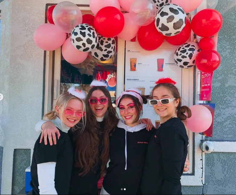 Group posing outside a cafe decorated with pink, red, and cow print balloons, wearing colorful sunglasses and festive headbands.