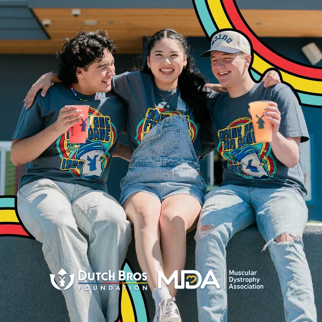 Three friends in matching blue t-shirts with drinks, sitting together outdoors. Dutch Bros Foundation and MDA logos below.