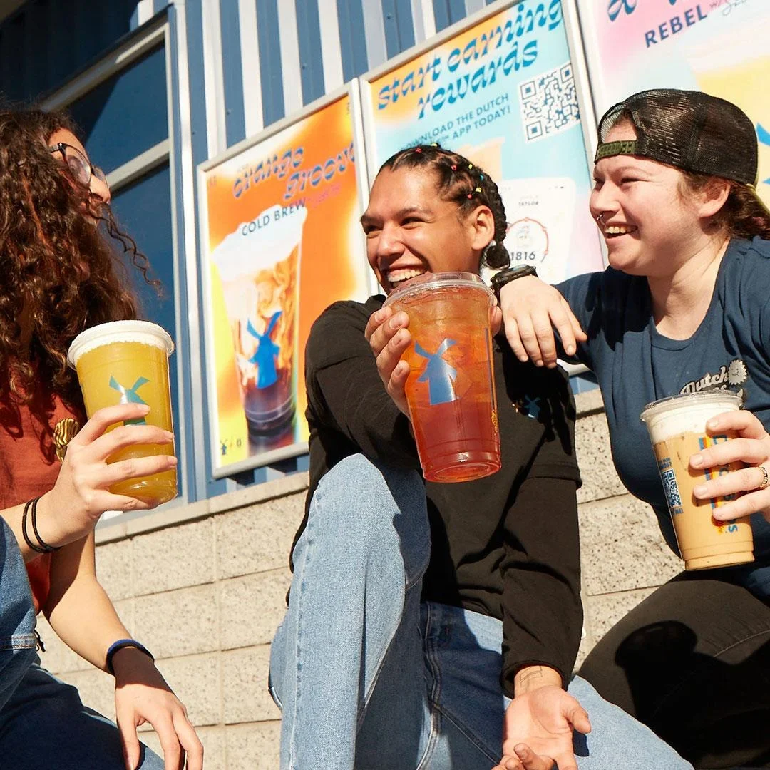 Three friends laughing and enjoying colorful cold brew drinks outside a Dutch Bros coffee shop on a sunny day.
