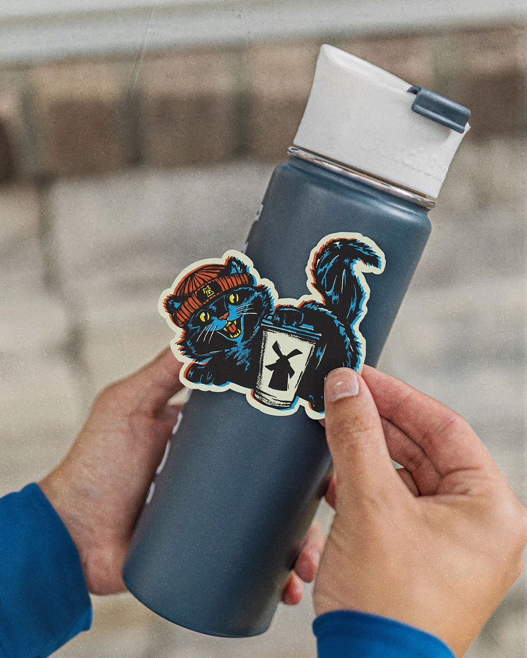 Hands applying a sticker of a black cat wearing an orange beanie to a dark blue insulated water bottle.