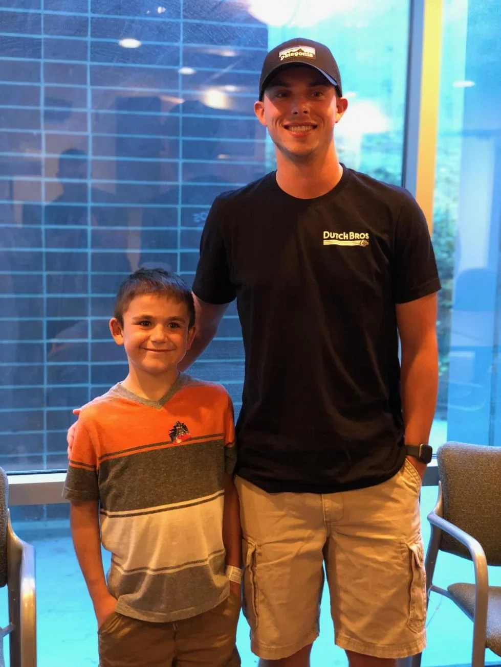 Dutch Bros employee in black shirt and cap posing with young boy in striped orange shirt against blue wall background.