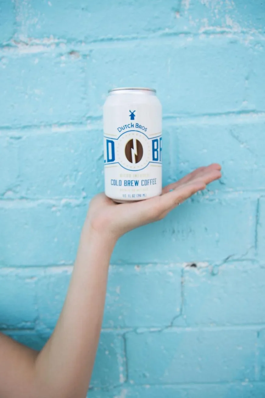 Hand holding a Dutch Bros cold brew coffee can against a turquoise blue brick wall.