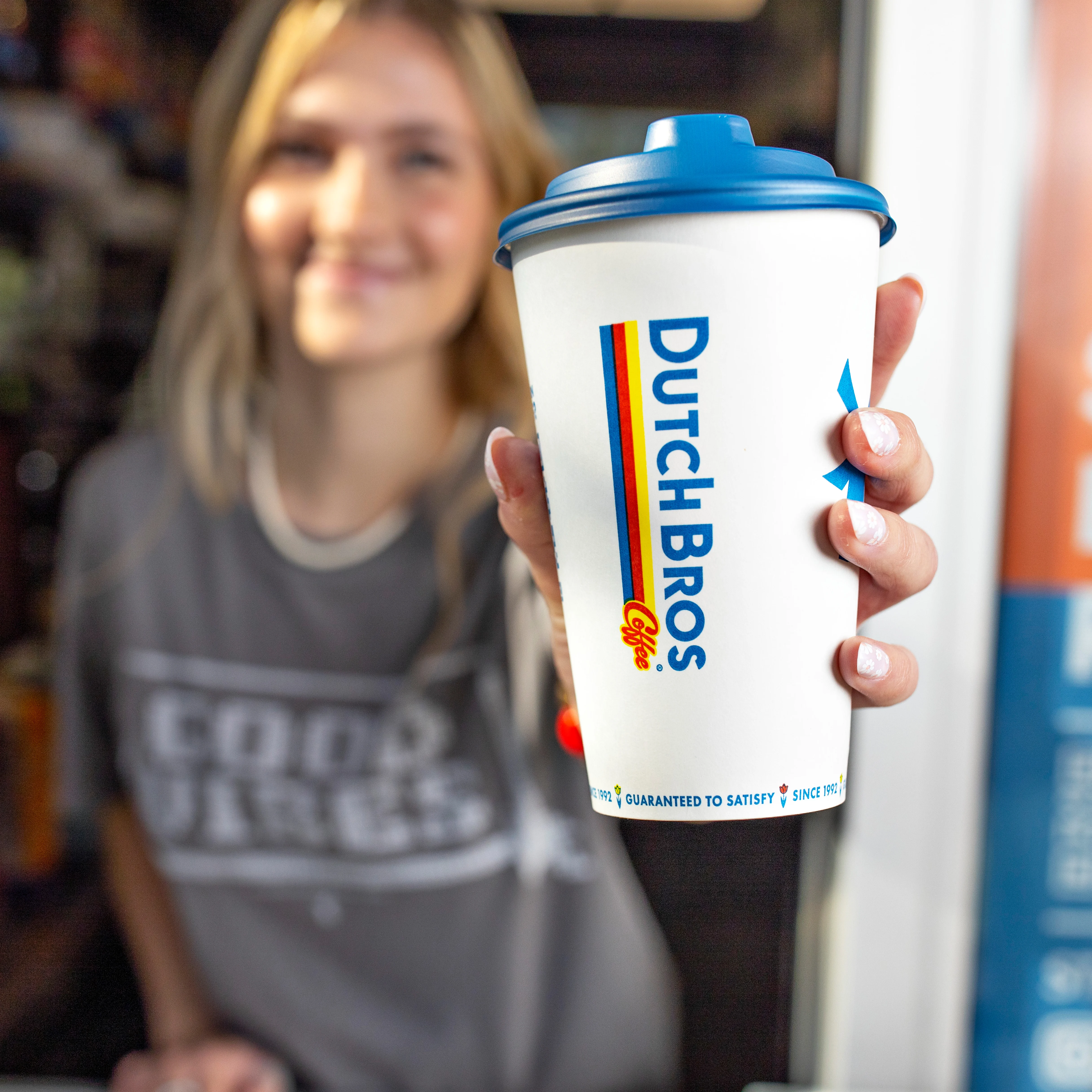 Hand holding a Dutch Bros coffee cup with blue lid, person wearing a gray shirt blurred in background.