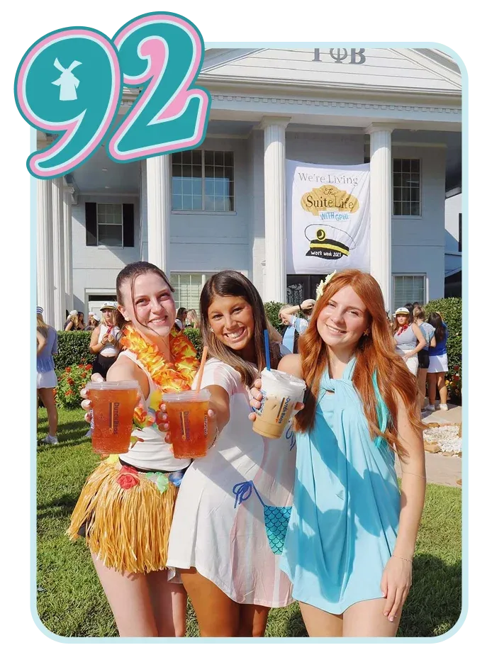 Three students celebrating at a sorority house with drinks, wearing themed outfits in front of a building.