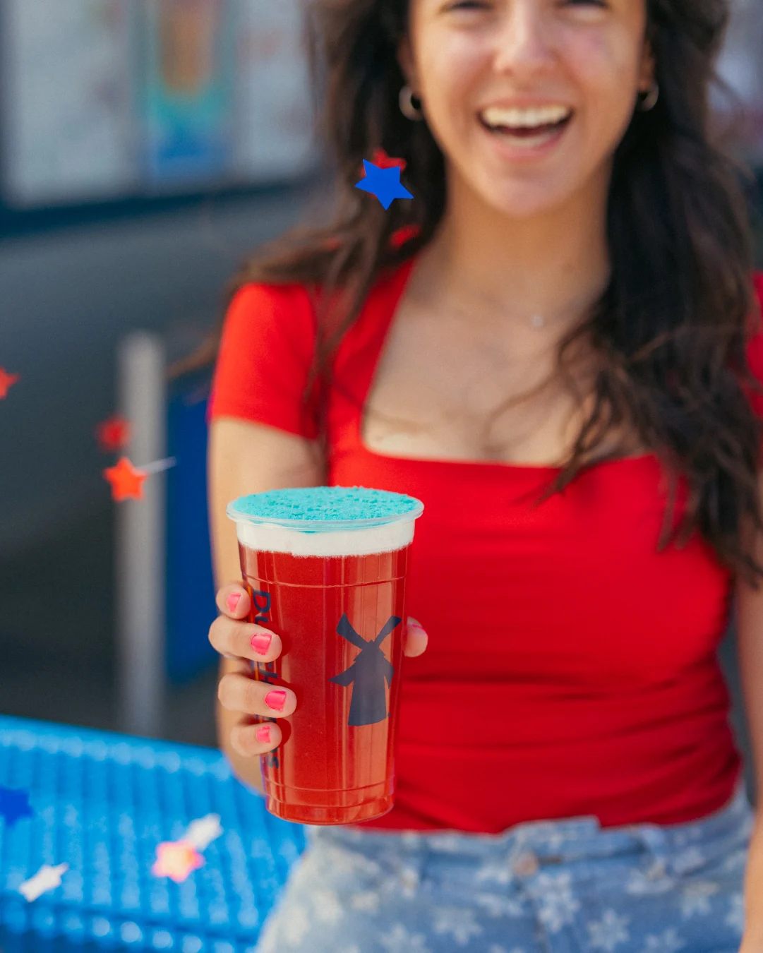 Person in red top holding a drink cup with blue lid and windmill design, decorated with colorful star graphics.