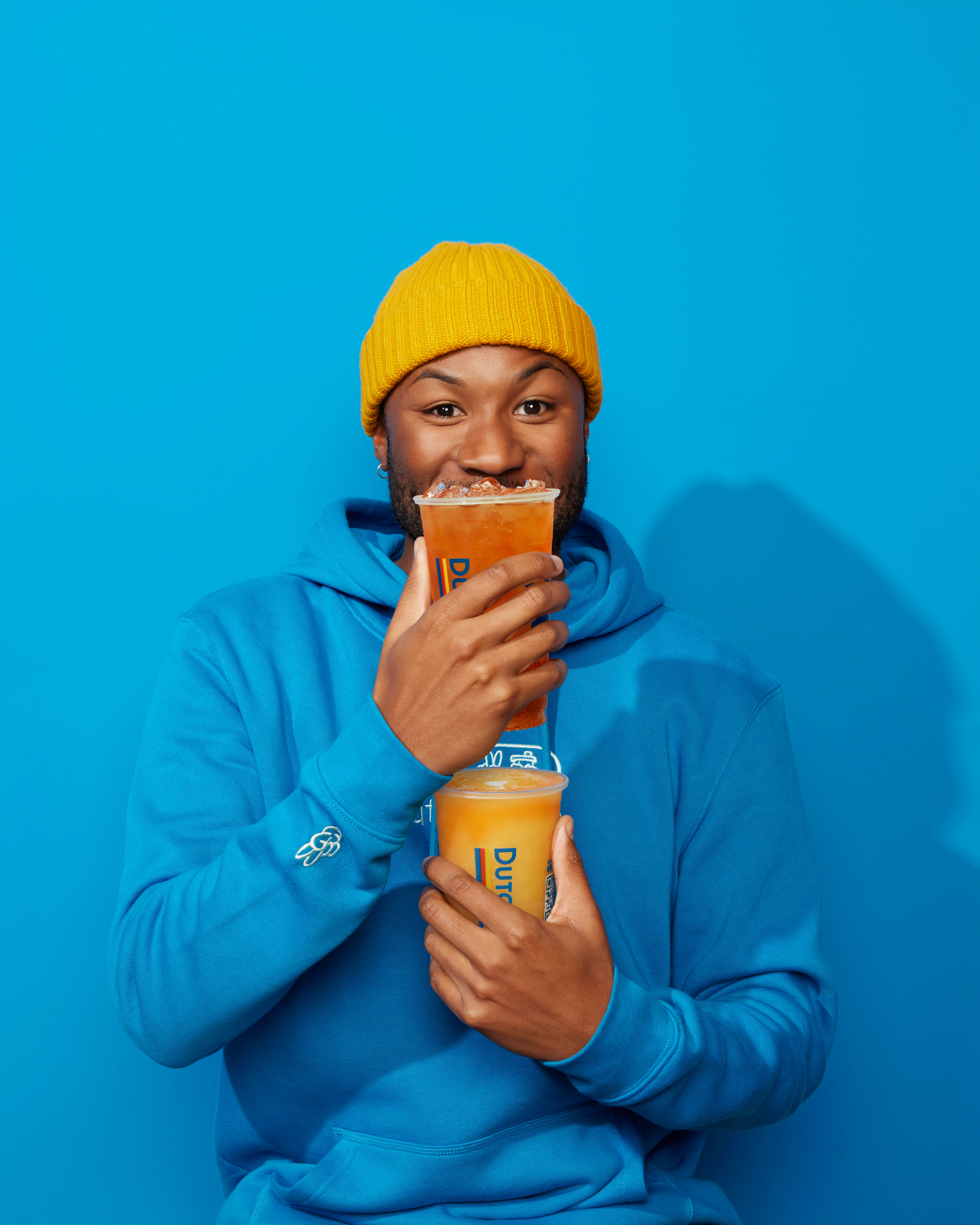 Person in bright yellow beanie and blue hoodie holding two orange drinks against vibrant blue background.