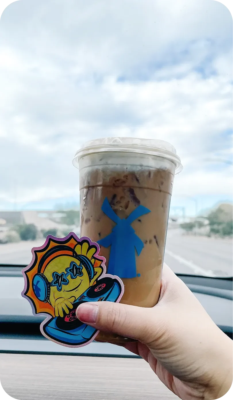 A hand holds an iced coffee cup with a blue windmill logo and a colorful DJ sun sticker, inside a car with a cloudy sky backdrop.