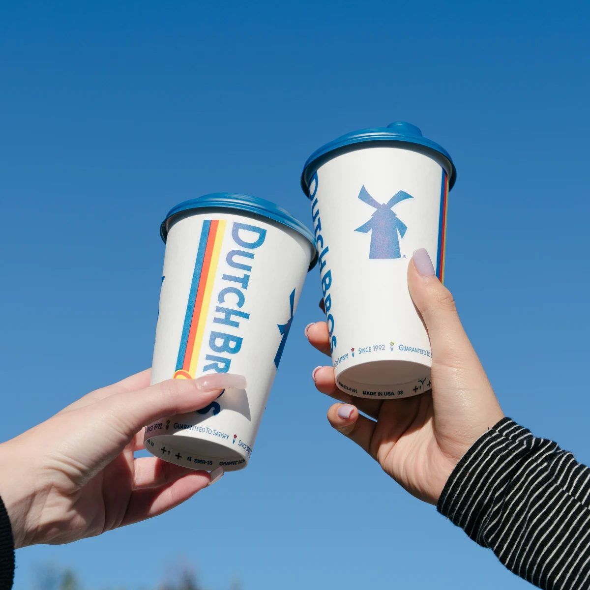 Two hands holding Dutch Bros coffee cups with blue lids against a bright blue sky, one cup featuring a windmill logo.