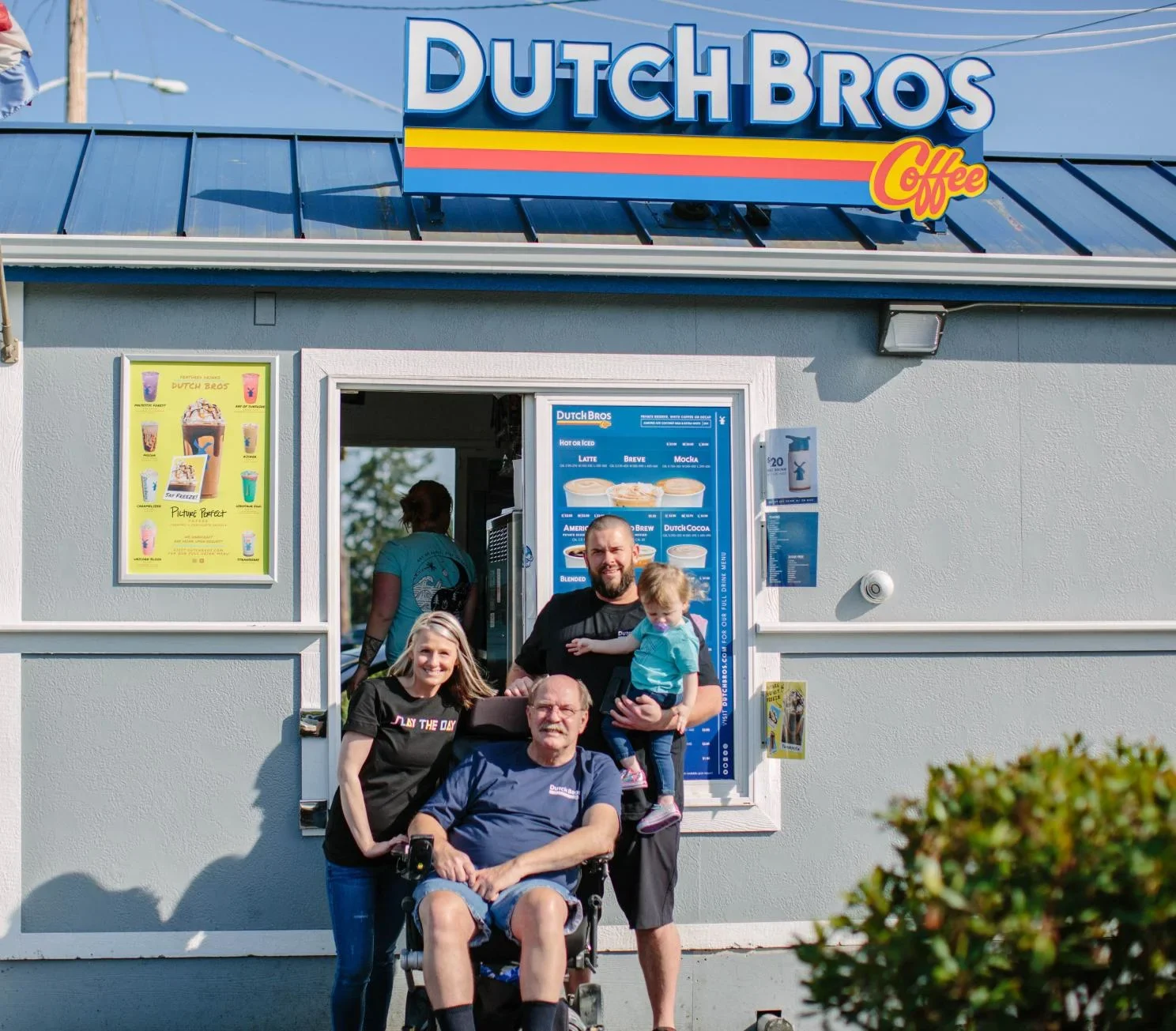 Family gathered outside a Dutch Bros Coffee shop with colorful signage and menu displays on a sunny day.