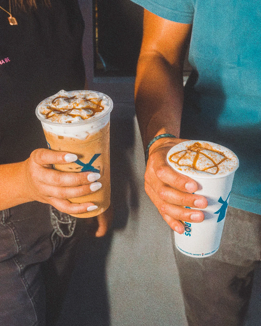 Two hands holding coffee drinks with caramel drizzle topping in branded cups with blue logo.
