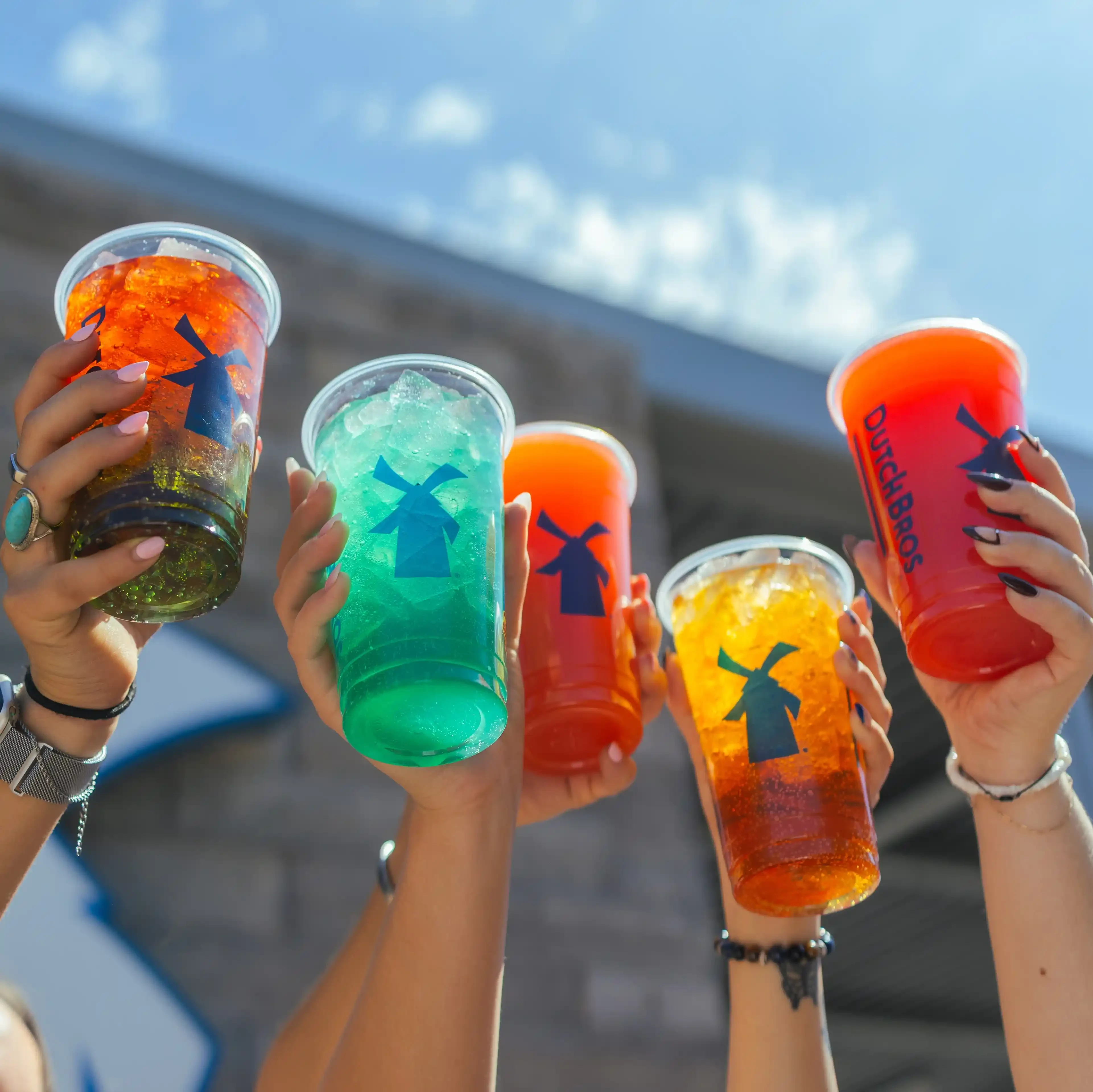 Four hands raised holding colorful Dutch Bros drinks in red, teal, orange, and yellow against a sunny blue sky.
