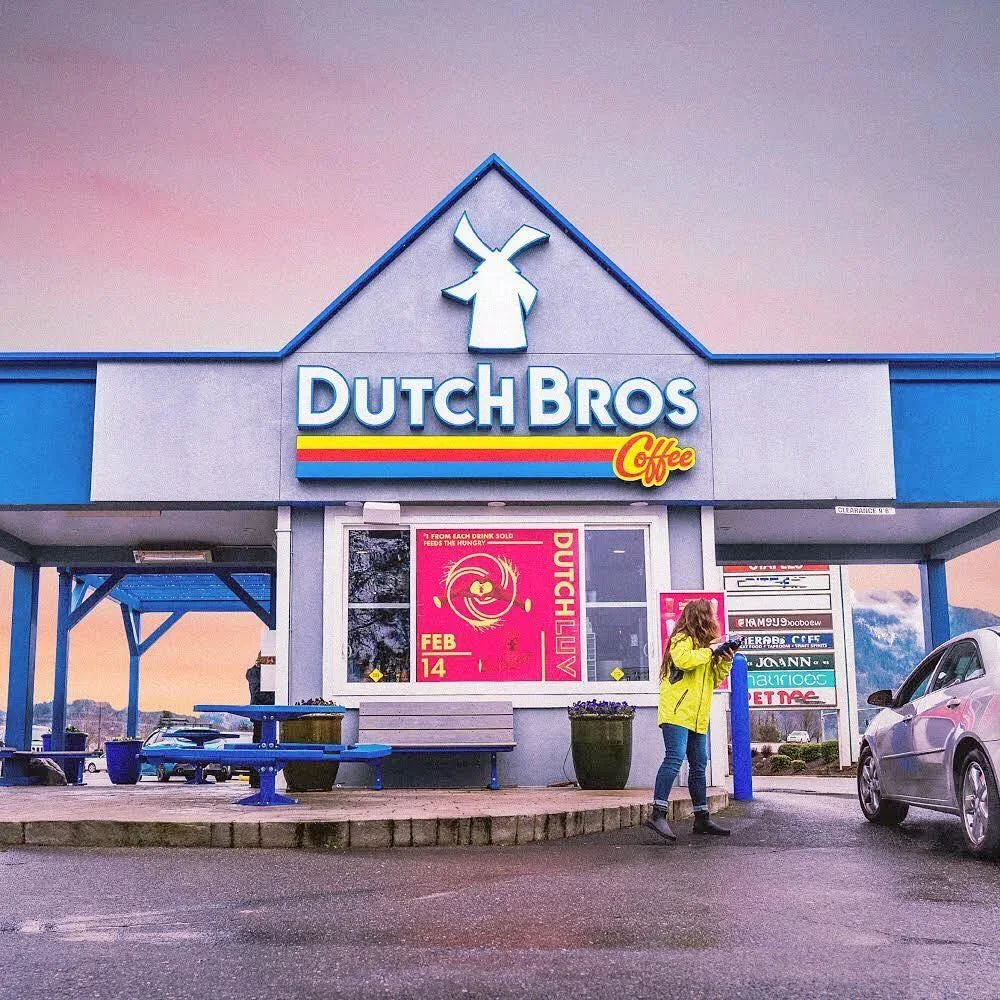 Dutch Bros Coffee storefront with windmill logo, blue trim and a person in yellow jacket standing outside at dusk.