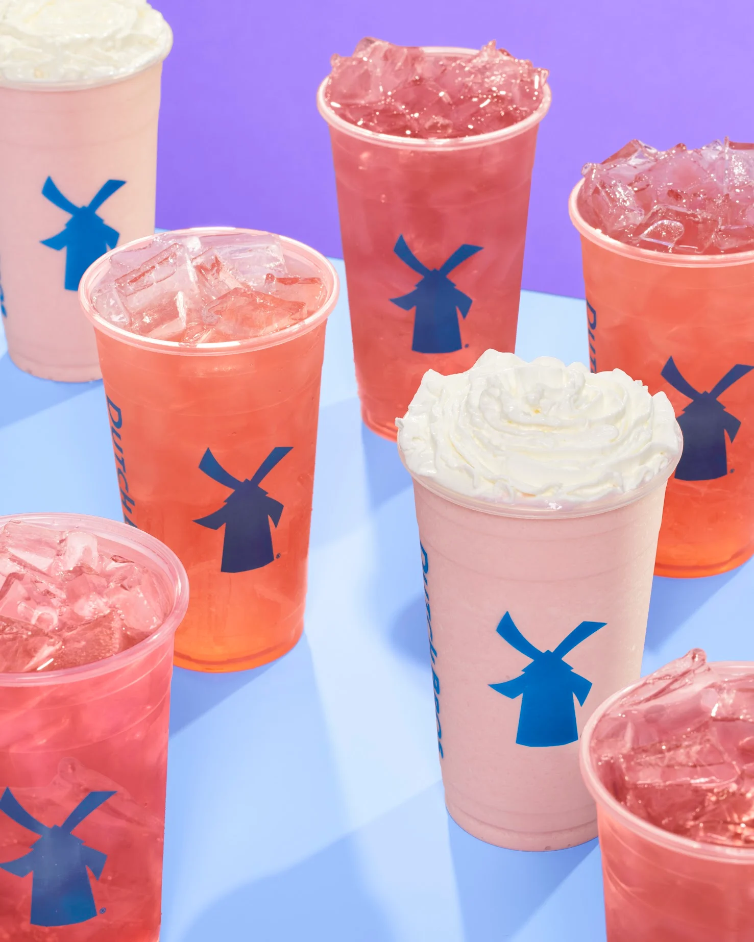 Colorful Dutch Bros drinks in clear cups with windmill logos - pink iced beverages and cream-topped shakes on blue background.