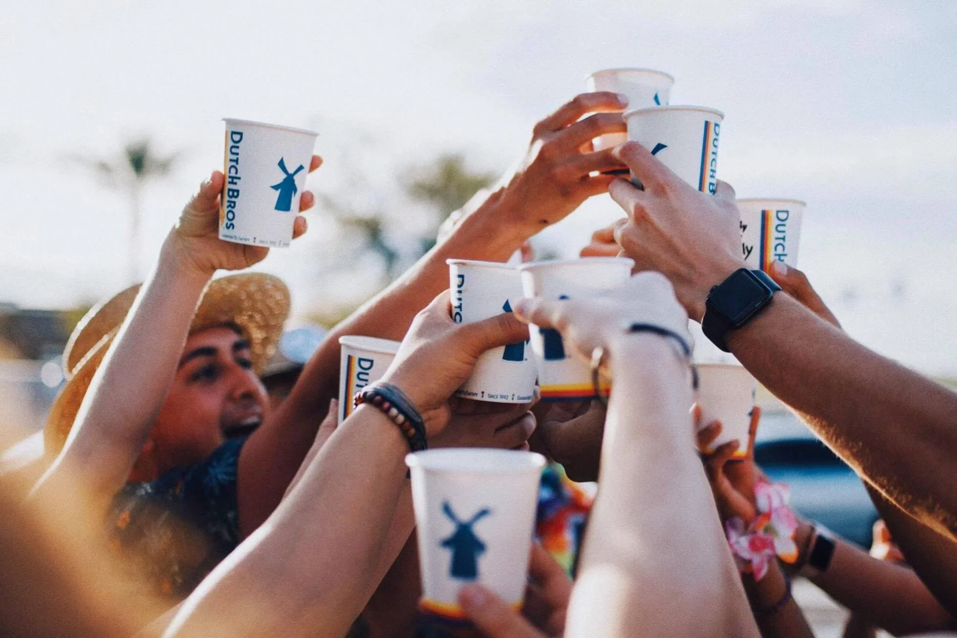 Hands raised in a toast with Dutch Bros coffee cups against a sunny outdoor backdrop with palm trees visible.