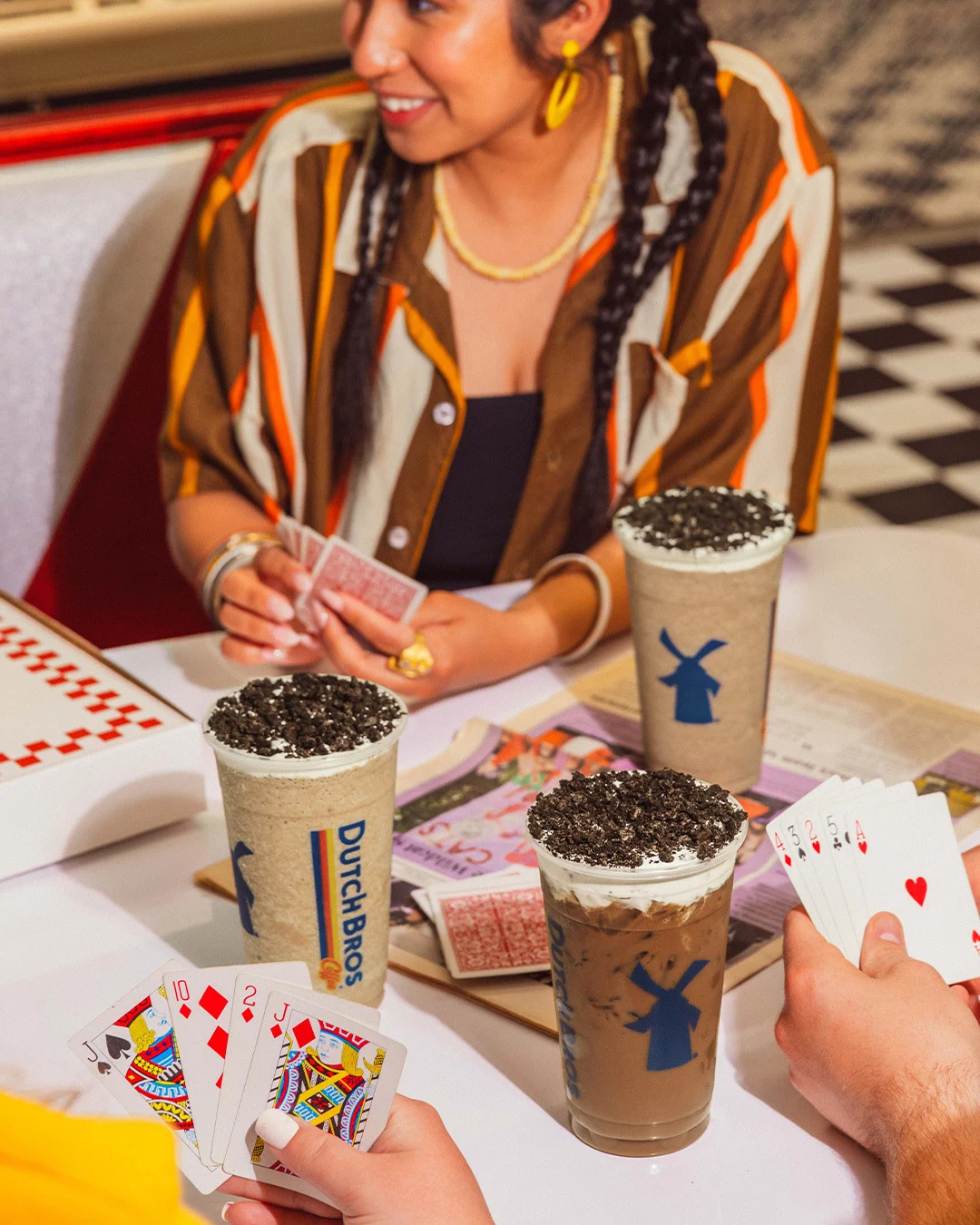 Person in striped shirt playing cards with friends at a café table with Dutch Bros coffee drinks topped with cookie crumbles.