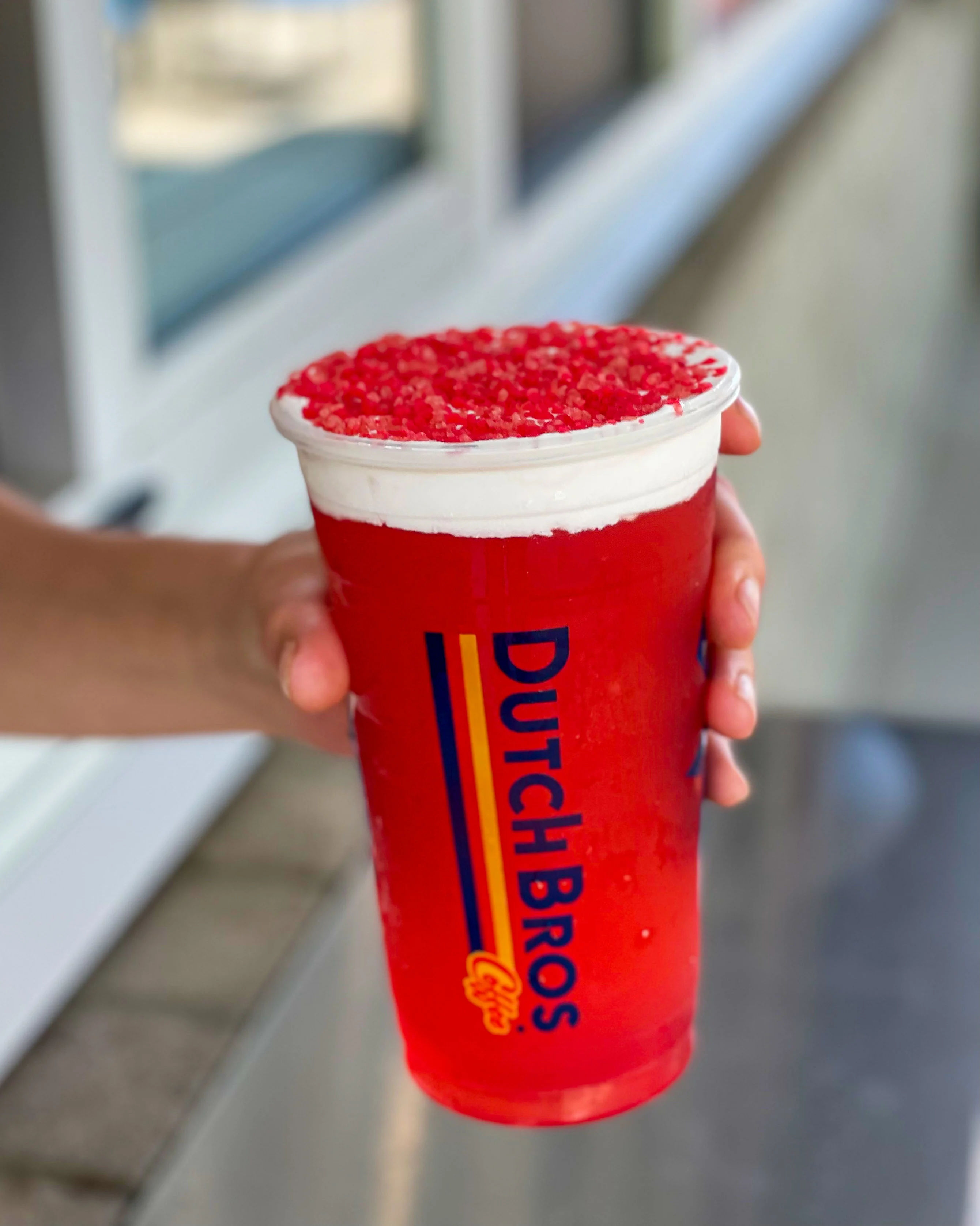 Hand holding a bright red Dutch Bros drink topped with white cream and red sprinkles in a branded cup.
