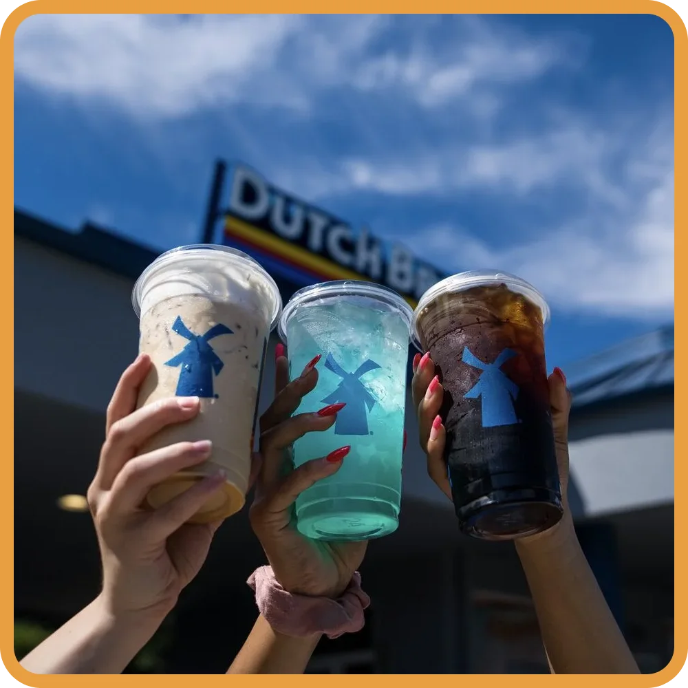 Three hands holding Dutch Bros drinks - a coffee, blue iced beverage, and dark soda - against blue sky background.