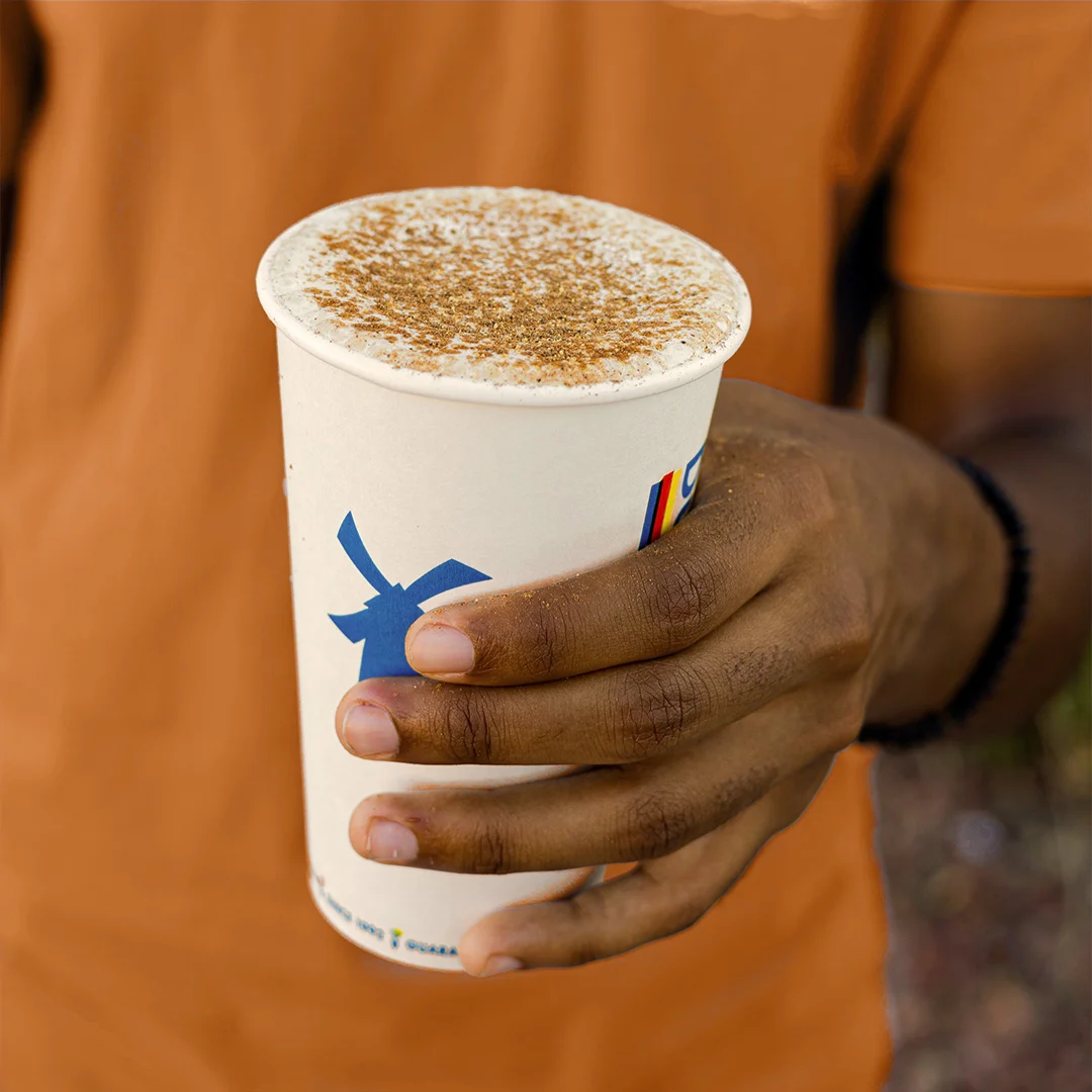 Hand holding a white coffee cup with blue windmill logo and cinnamon-sprinkled foam on top.