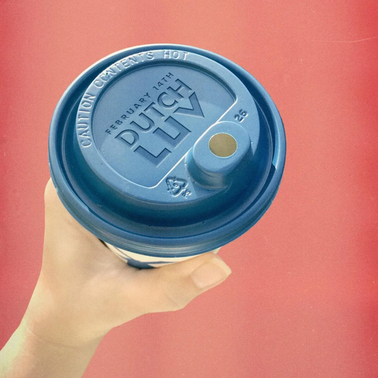 Hand holding coffee cup with blue lid marked "DUTCH" for February 14th event against coral pink background.