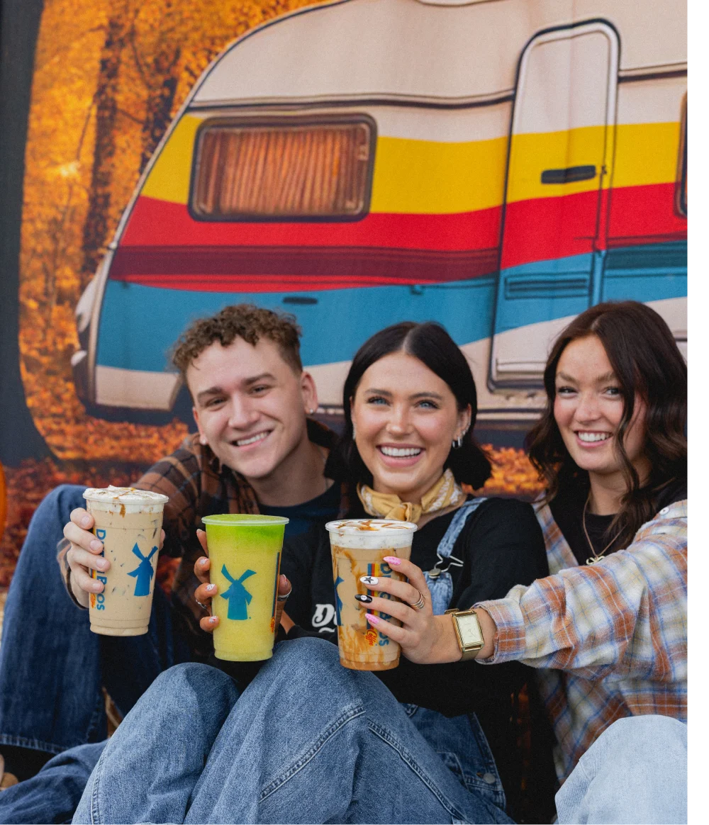 Three smiling friends holding Dutch Bros drinks in front of a colorful vintage camper with autumn foliage in the background.