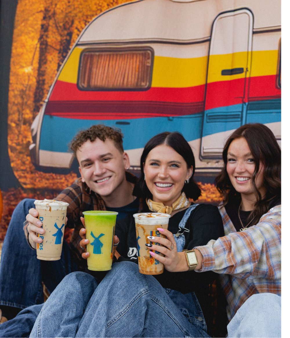 Three smiling friends holding Dutch Bros drinks in front of a colorful vintage camper with autumn foliage in the background.