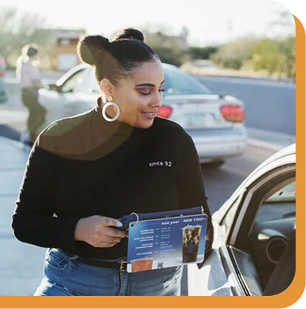 Woman with hair bun and hoop earrings wearing black top and jeans, holding an ipad next to a parked car.