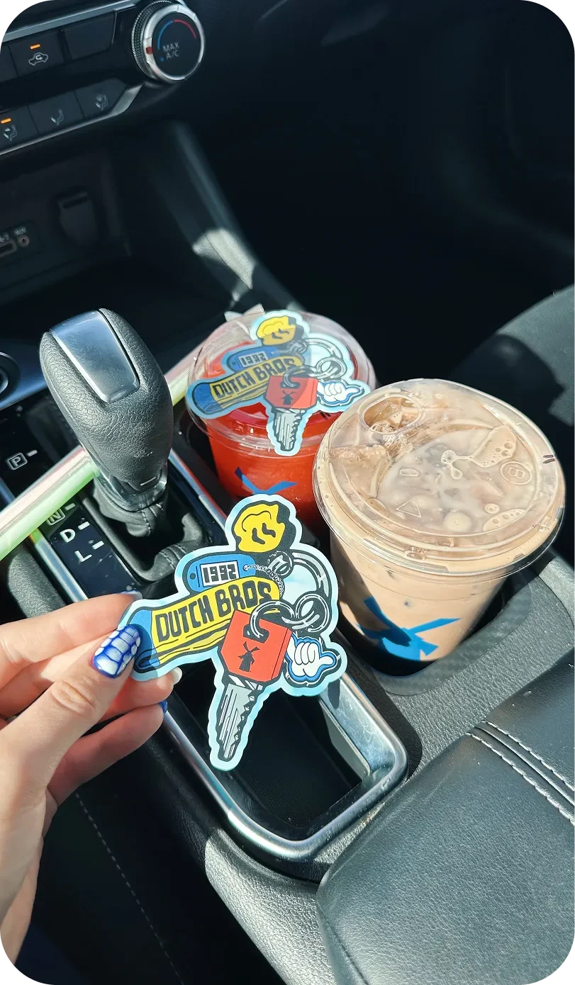 A hand holding a Dutch Bros sticker inside a car, with two Dutch Bros iced drinks in the center console cup holders.