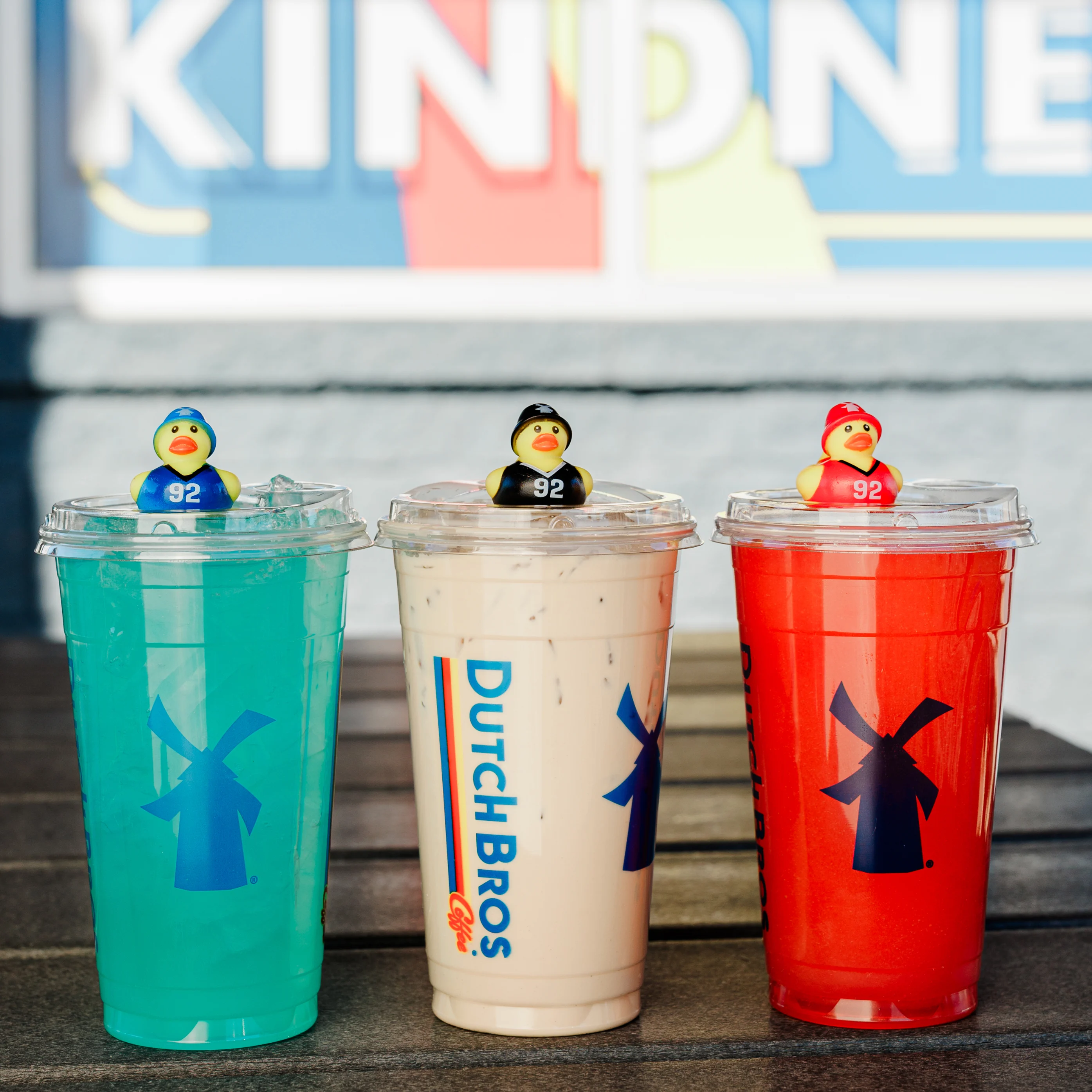 Three Dutch Bros drinks in blue, tan, and red cups, each topped with a colorful rubber duck wearing number 92.