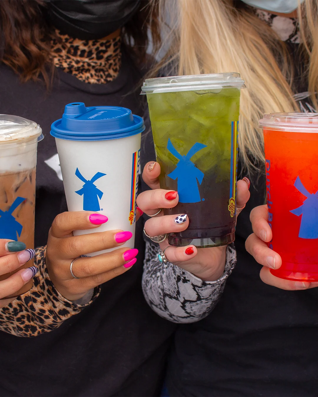 Hands with colorful manicures holding Dutch Bros coffee drinks with blue windmill logos in white, green, and red cups.