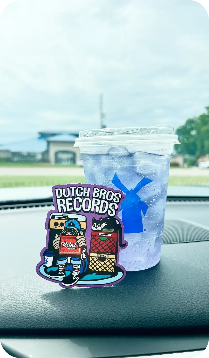 A Dutch Bros iced drink on a car dashboard beside a colorful Dutch Bros Records sticker featuring cartoon characters.