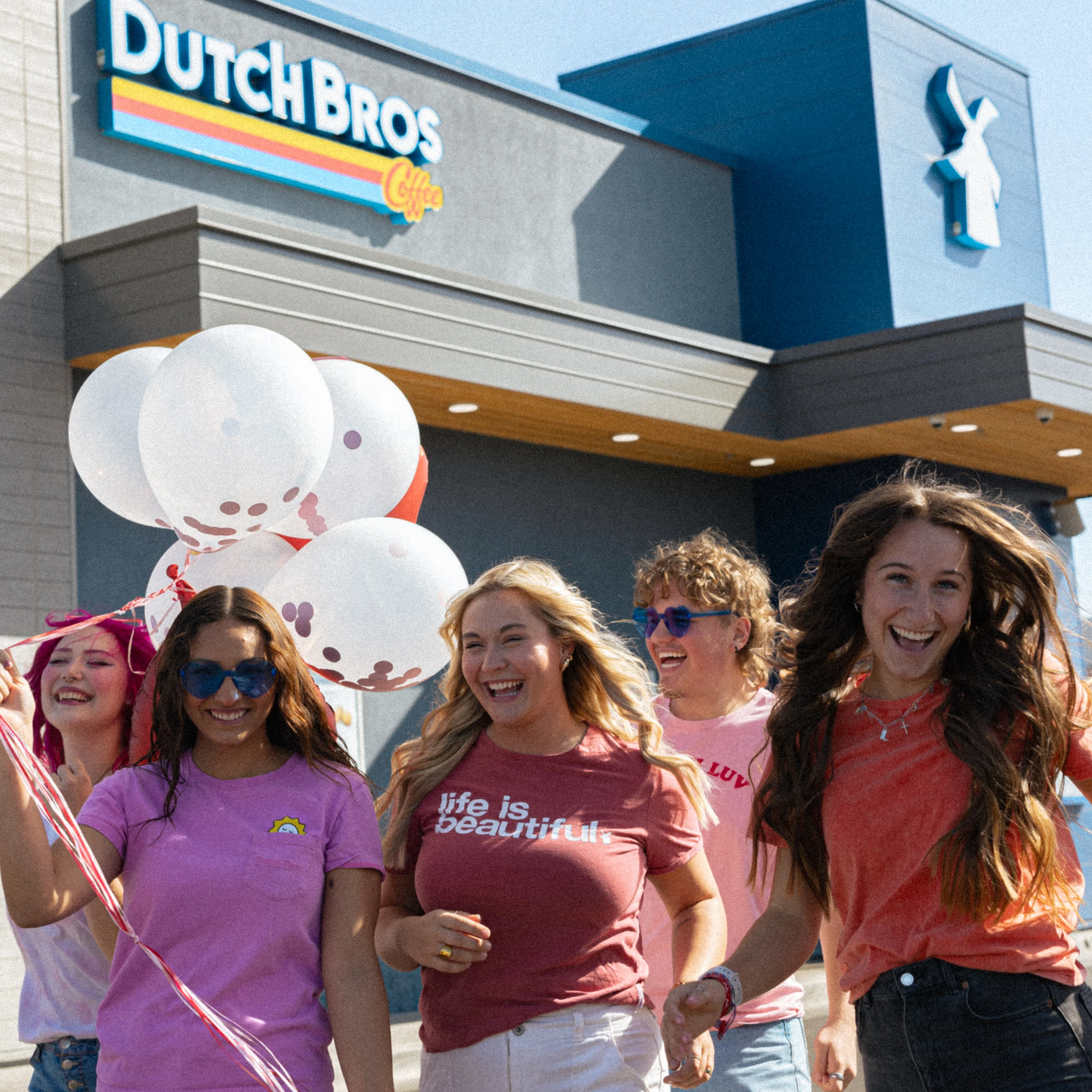 Group of smiling friends in colorful shirts with white balloons outside a Dutch Bros Coffee shop.
