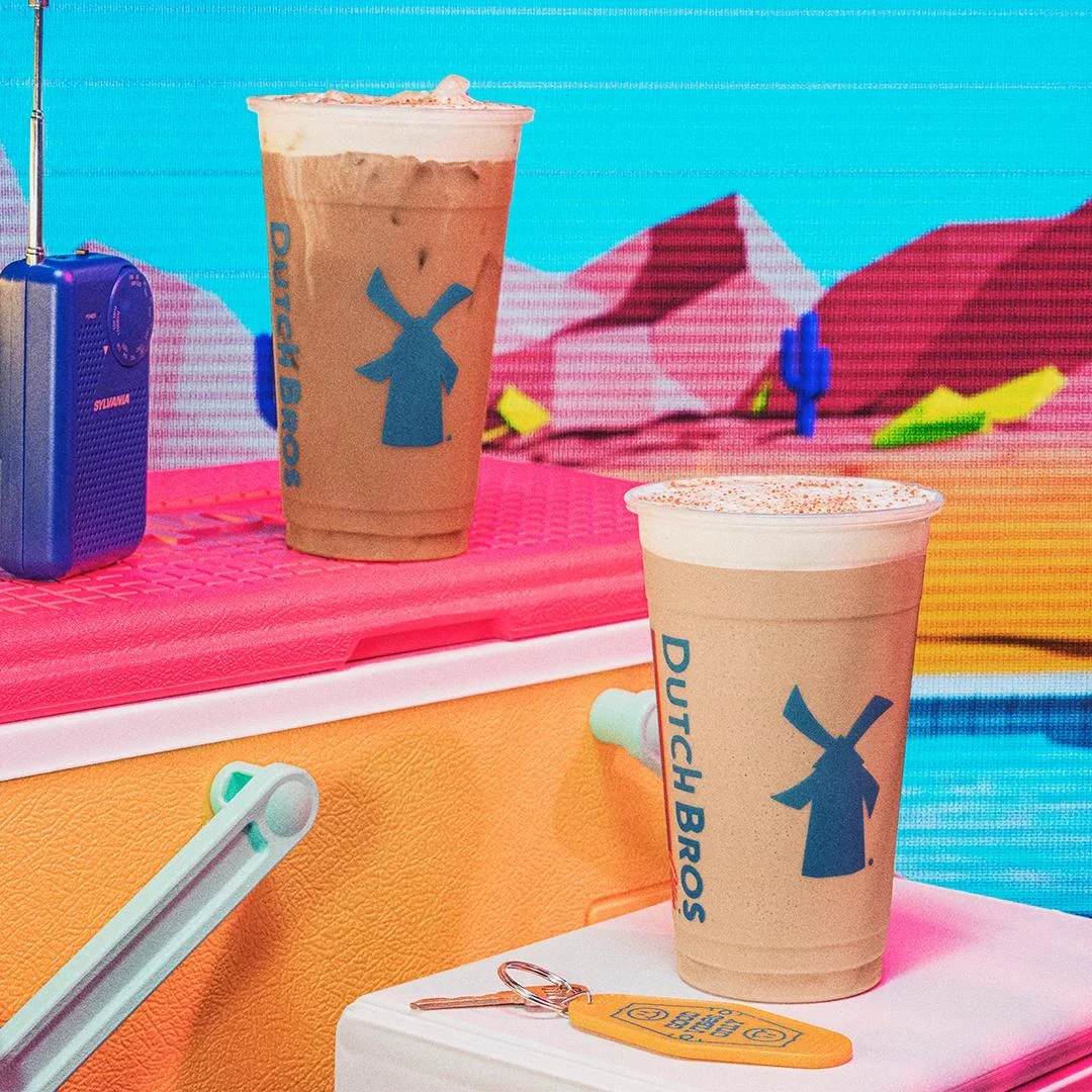 Two Dutch Bros iced coffee drinks with blue windmill logos against a colorful desert-themed background with a radio.