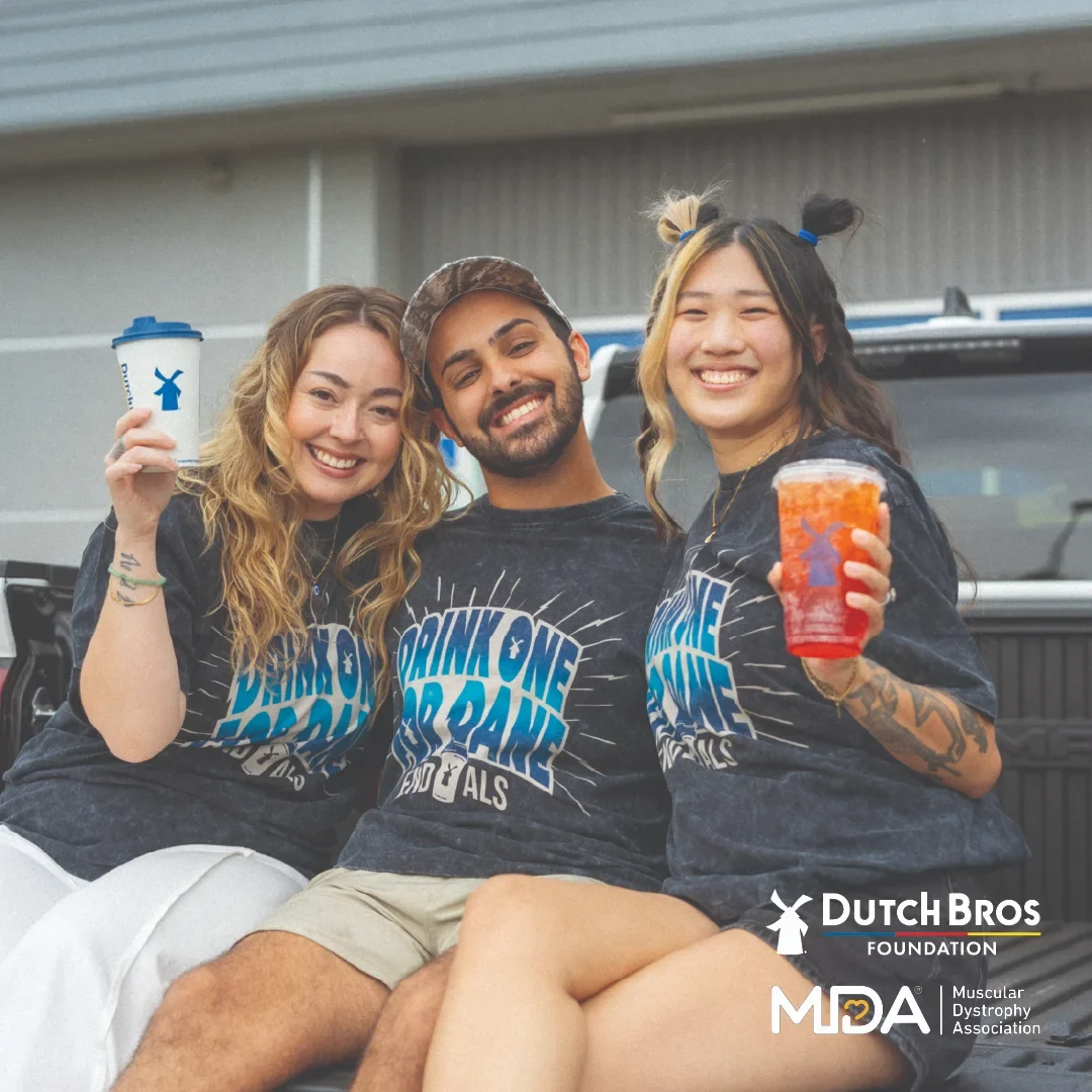 Three smiling friends in Dutch Bros "Drink One for Dane" ALS shirts holding beverages, with Dutch Bros Foundation and MDA logos.