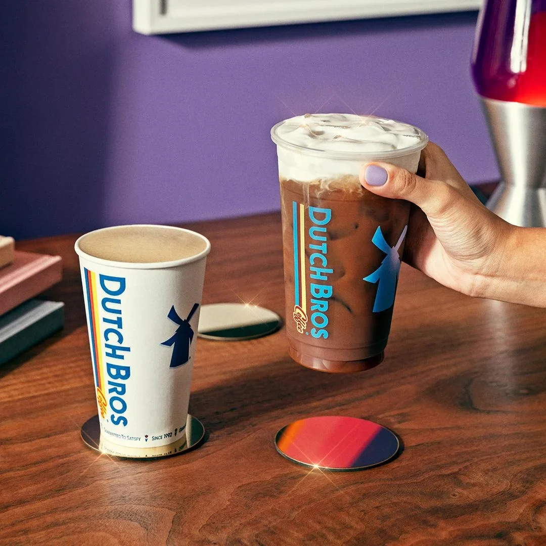 Hand holding iced Dutch Bros coffee drink next to a hot beverage cup on wooden table with purple wall background.