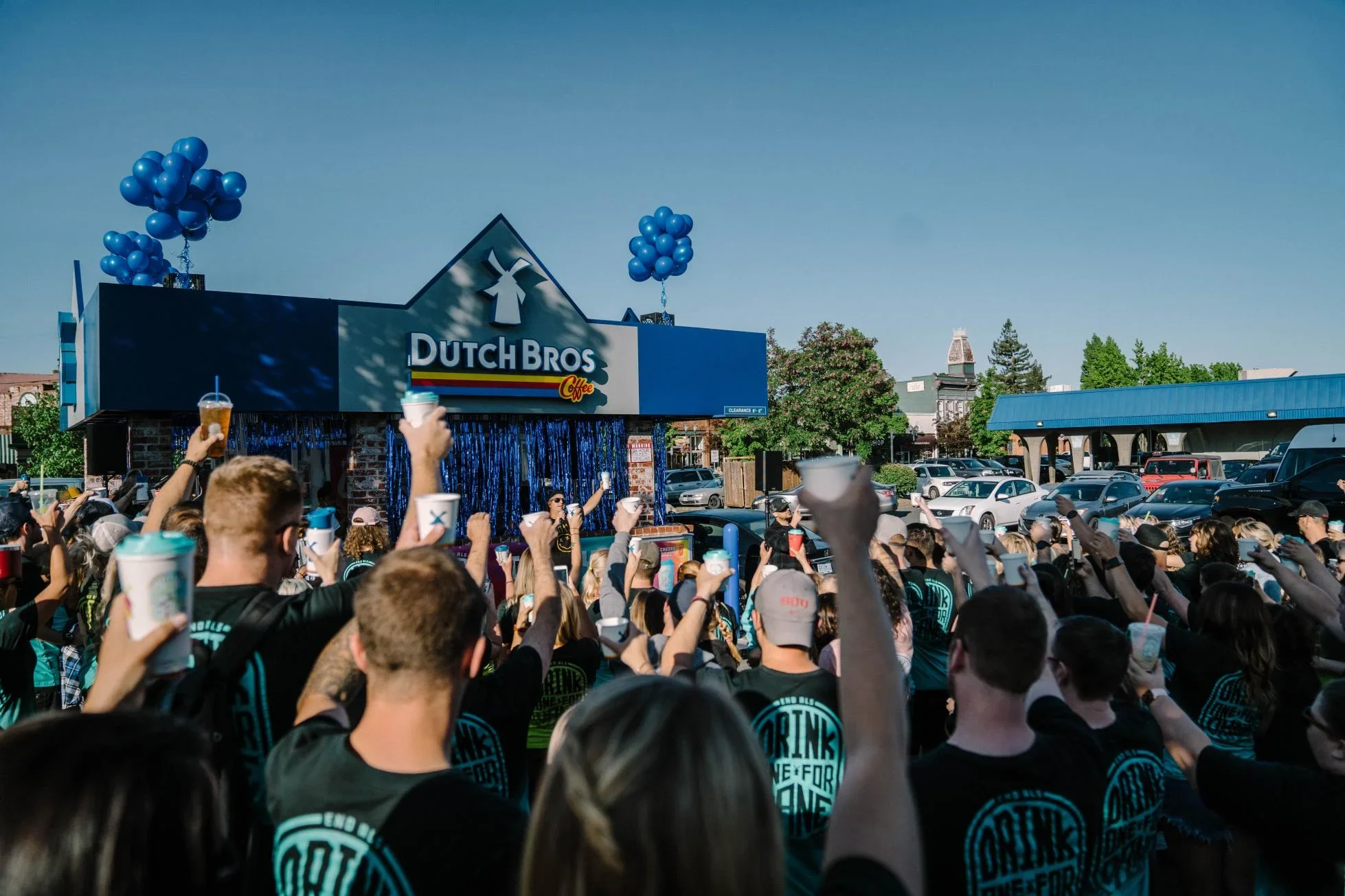 Crowd raising drinks at Dutch Bros Coffee grand opening celebration with blue balloons decorating the storefront.
