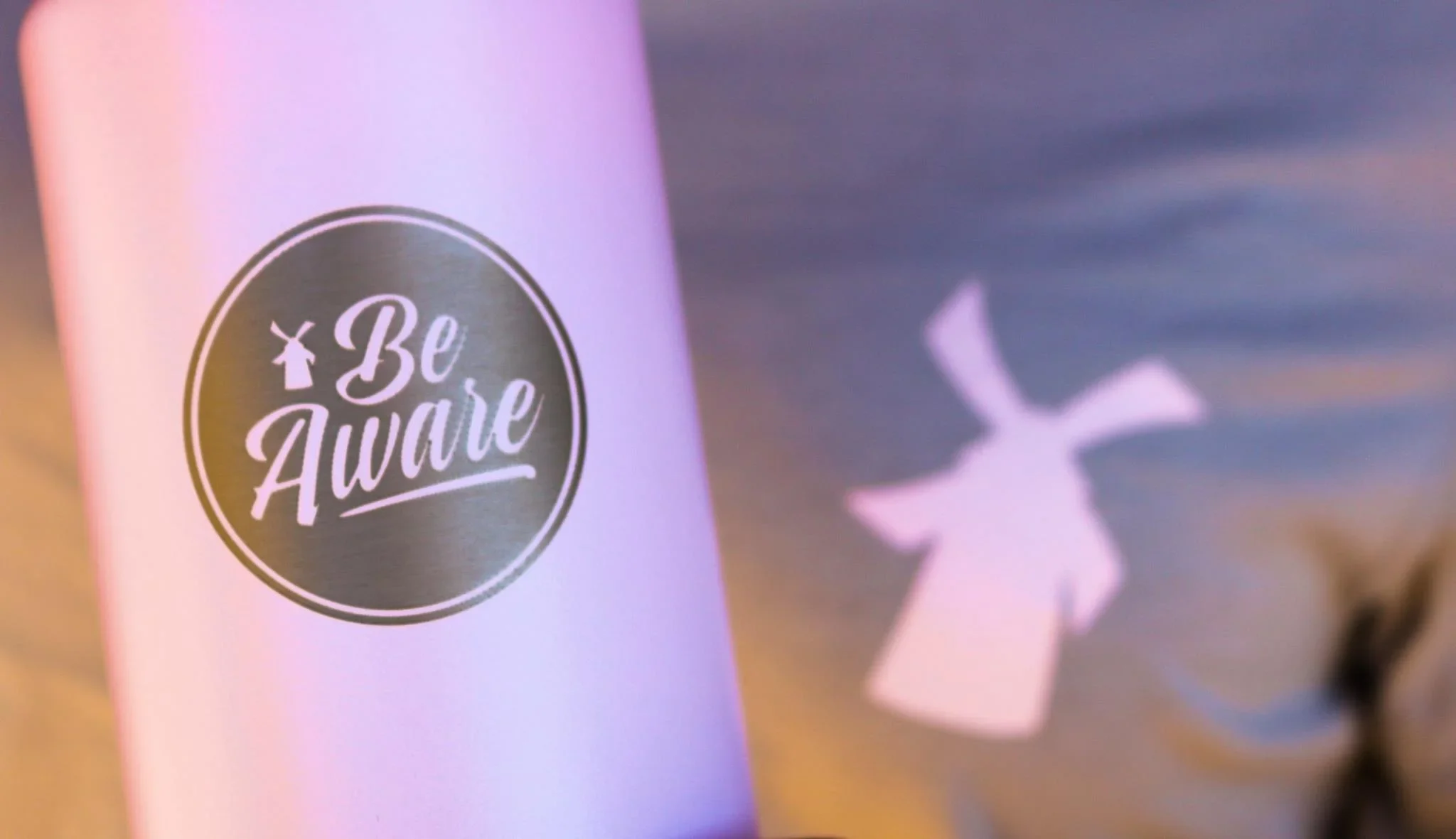 Pink tumbler with "Be Aware" logo featuring a windmill silhouette, with a blurred windmill shape in the background.