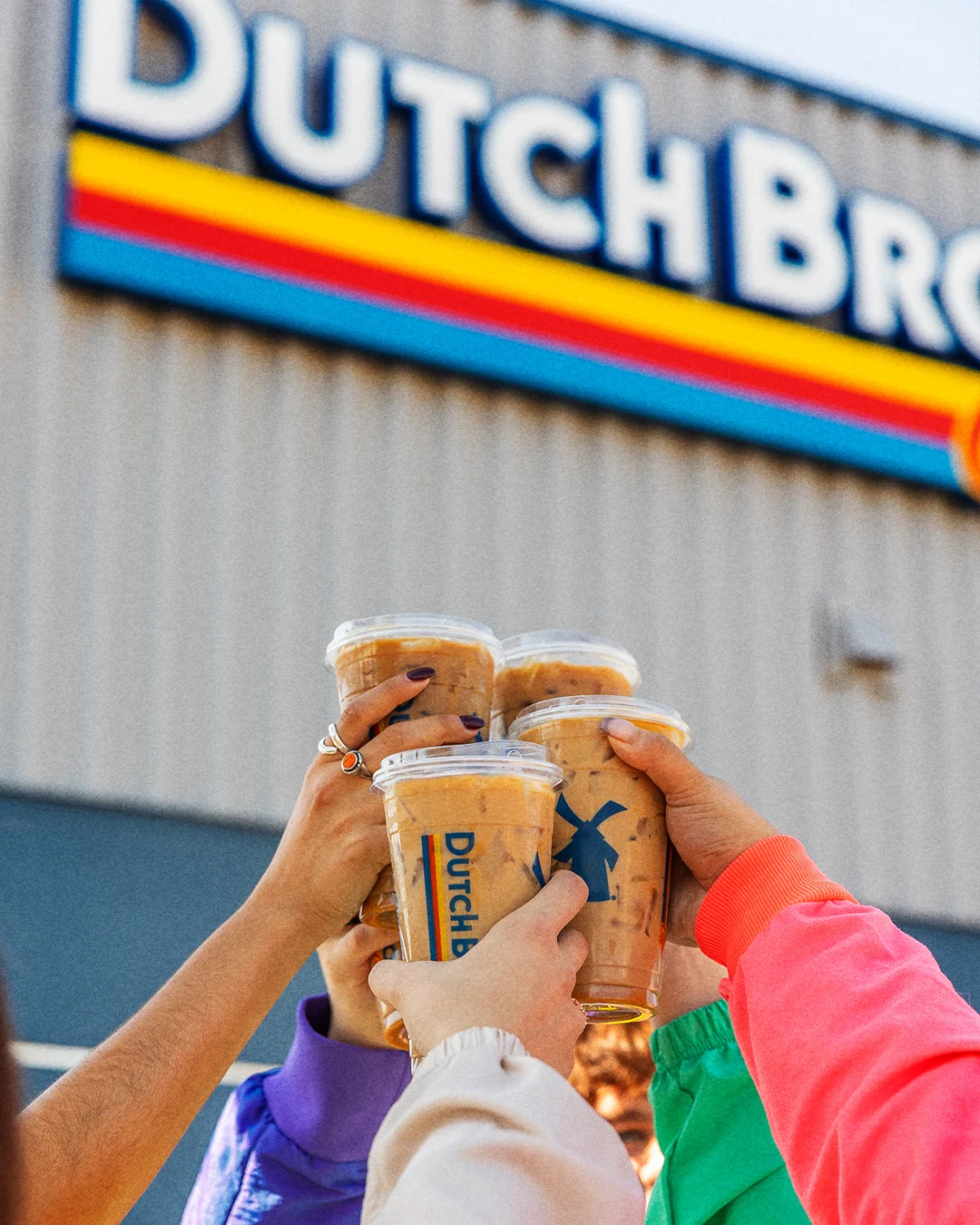 Hands in colorful sleeves toasting with Dutch Bros coffee drinks outside the store with its distinctive logo visible.