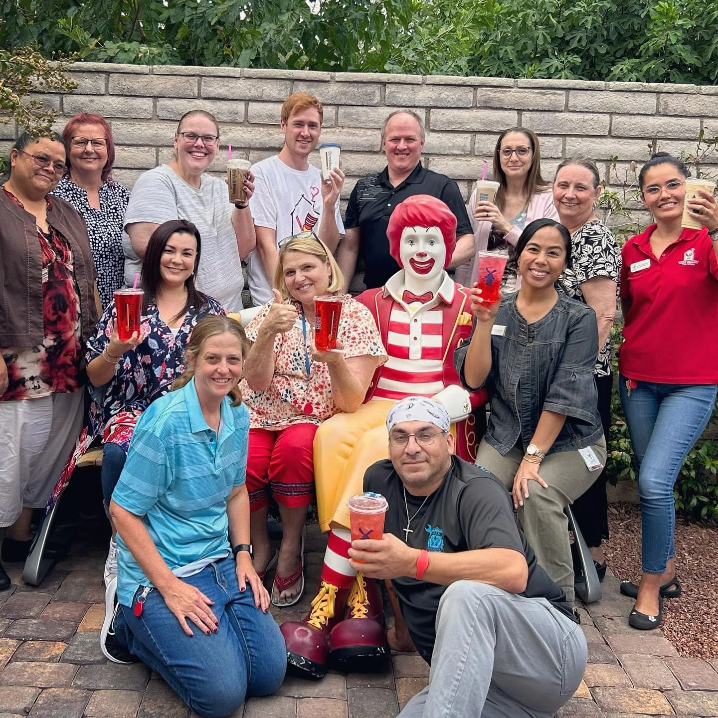 Group gathered around Ronald McDonald statue at outdoor event, everyone holding colorful drinks and smiling for photo.