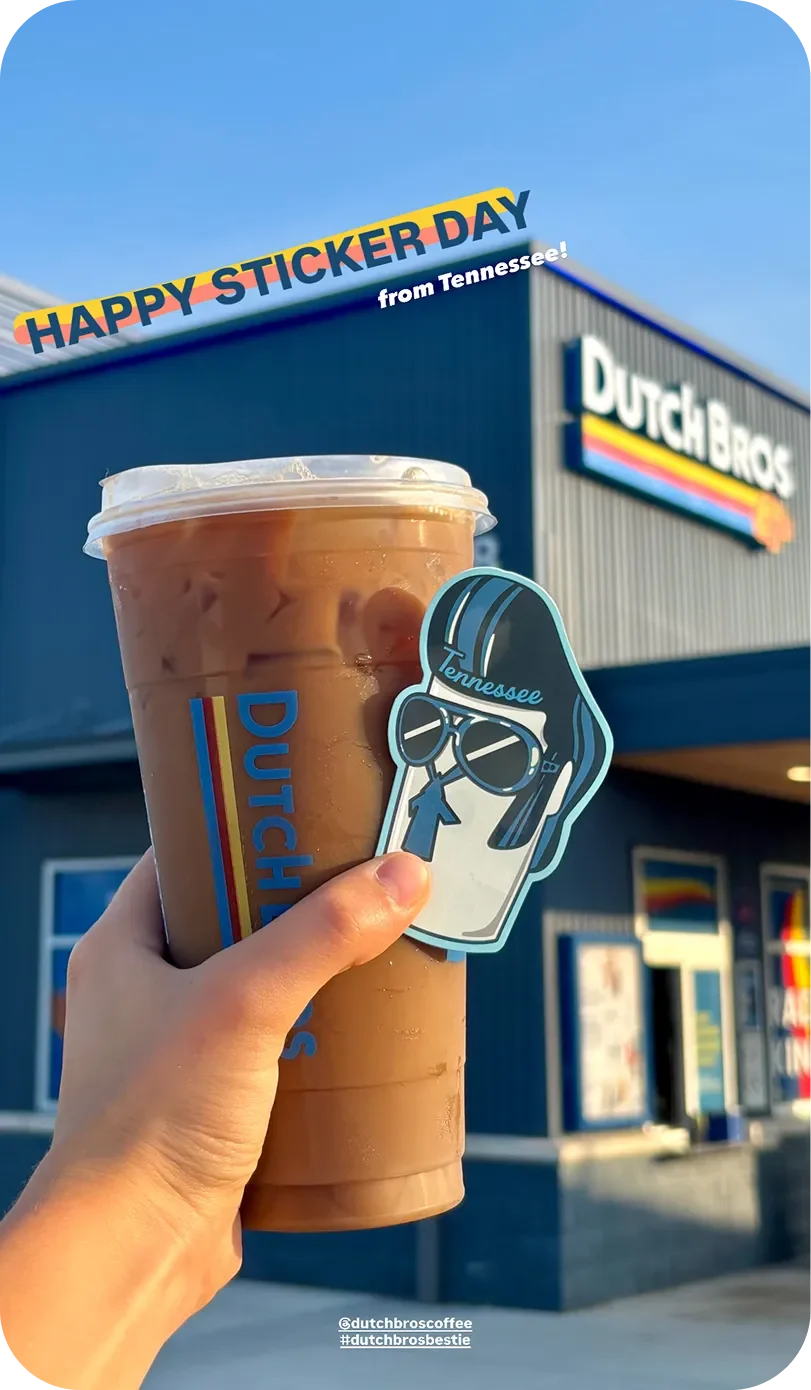 A hand holds a Dutch Bros iced coffee and a Tennessee sticker in front of a Dutch Bros store on Sticker Day.