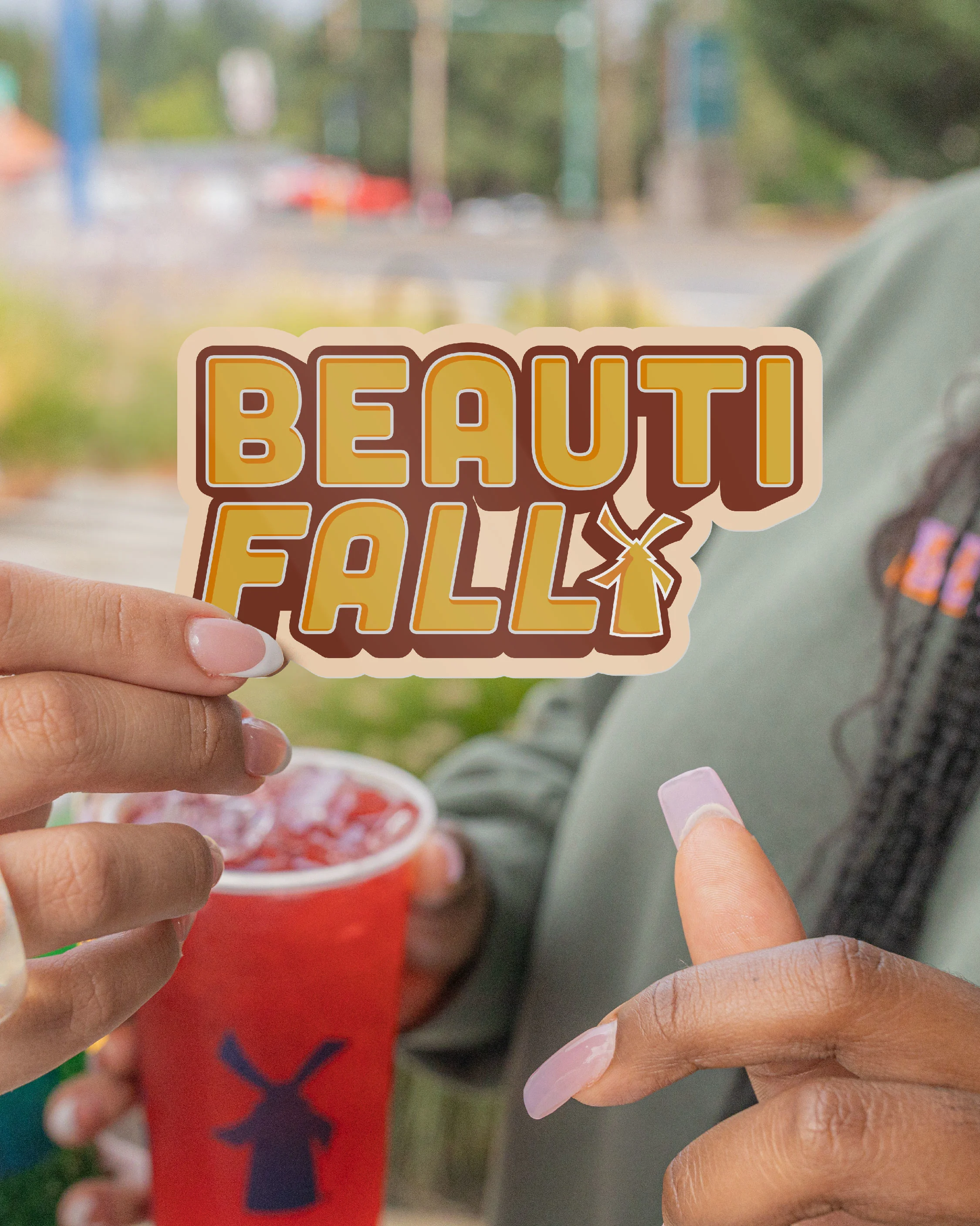 Hands holding a yellow "BEAUTI FALL" sticker with windmill icon and a red drink cup featuring a windmill silhouette.